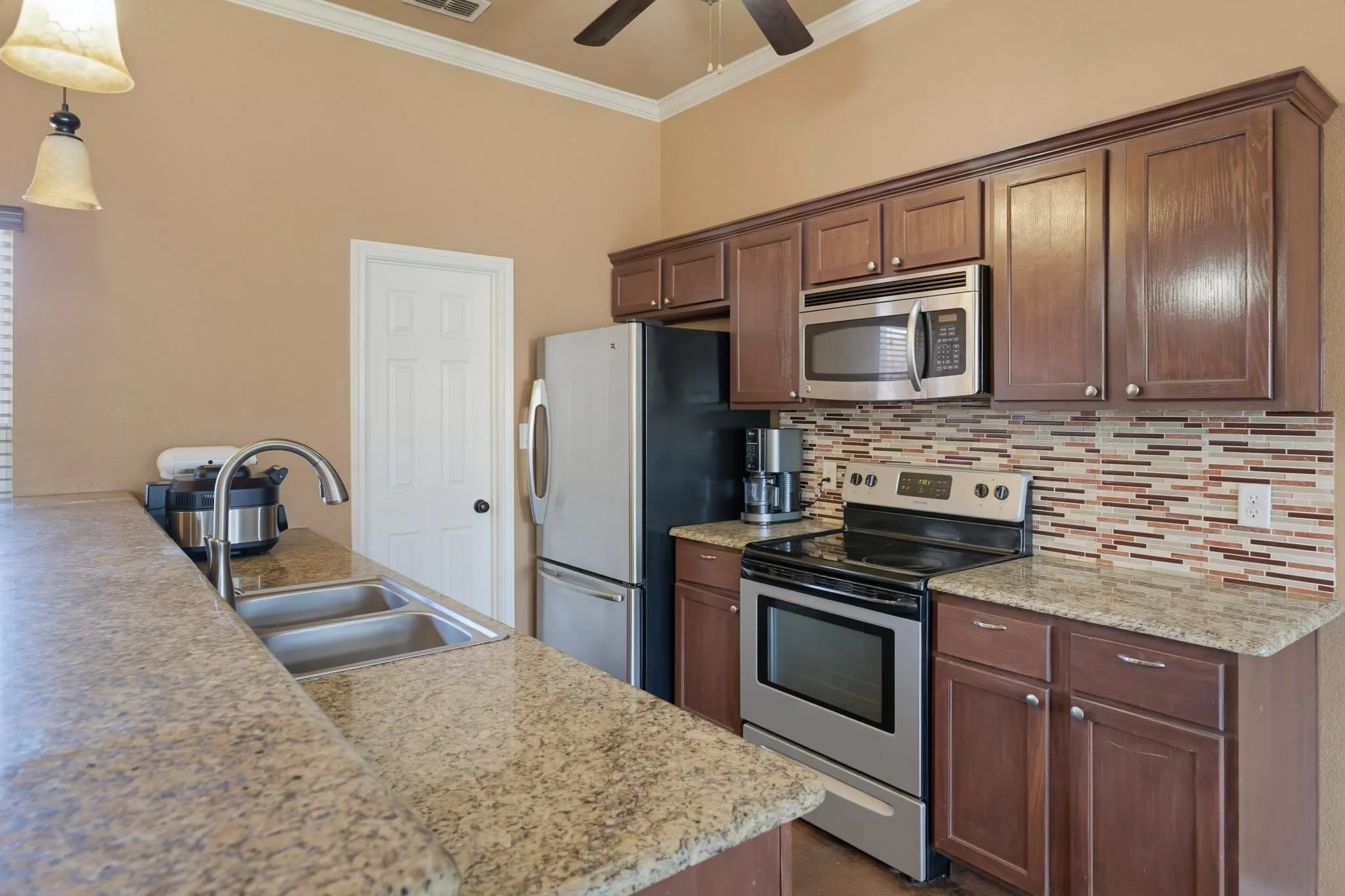 Kitchen featuring wood-finish cabinetry, light-toned countertops, a stainless steel double basin sink, a patterned tile backsplash, and stainless steel appliances