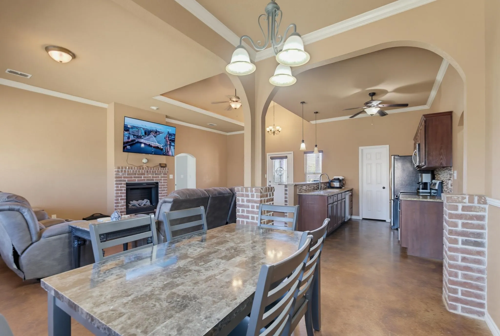 Open-concept living space featuring a brick fireplace surround, arched doorways, a tray ceiling, and a kitchen with dark wood-finish cabinetry