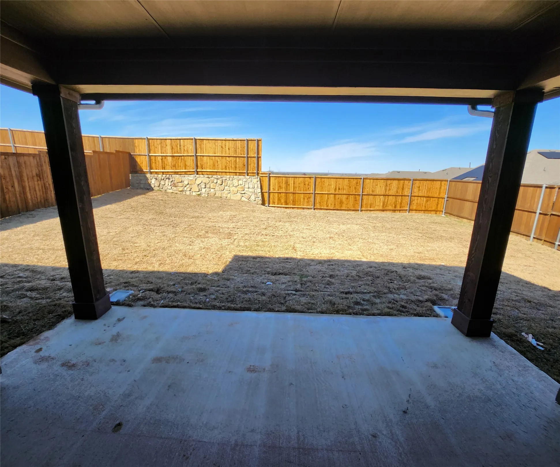 Covered concrete patio opening to an expansive backyard with wood fencing and a stone retaining wall