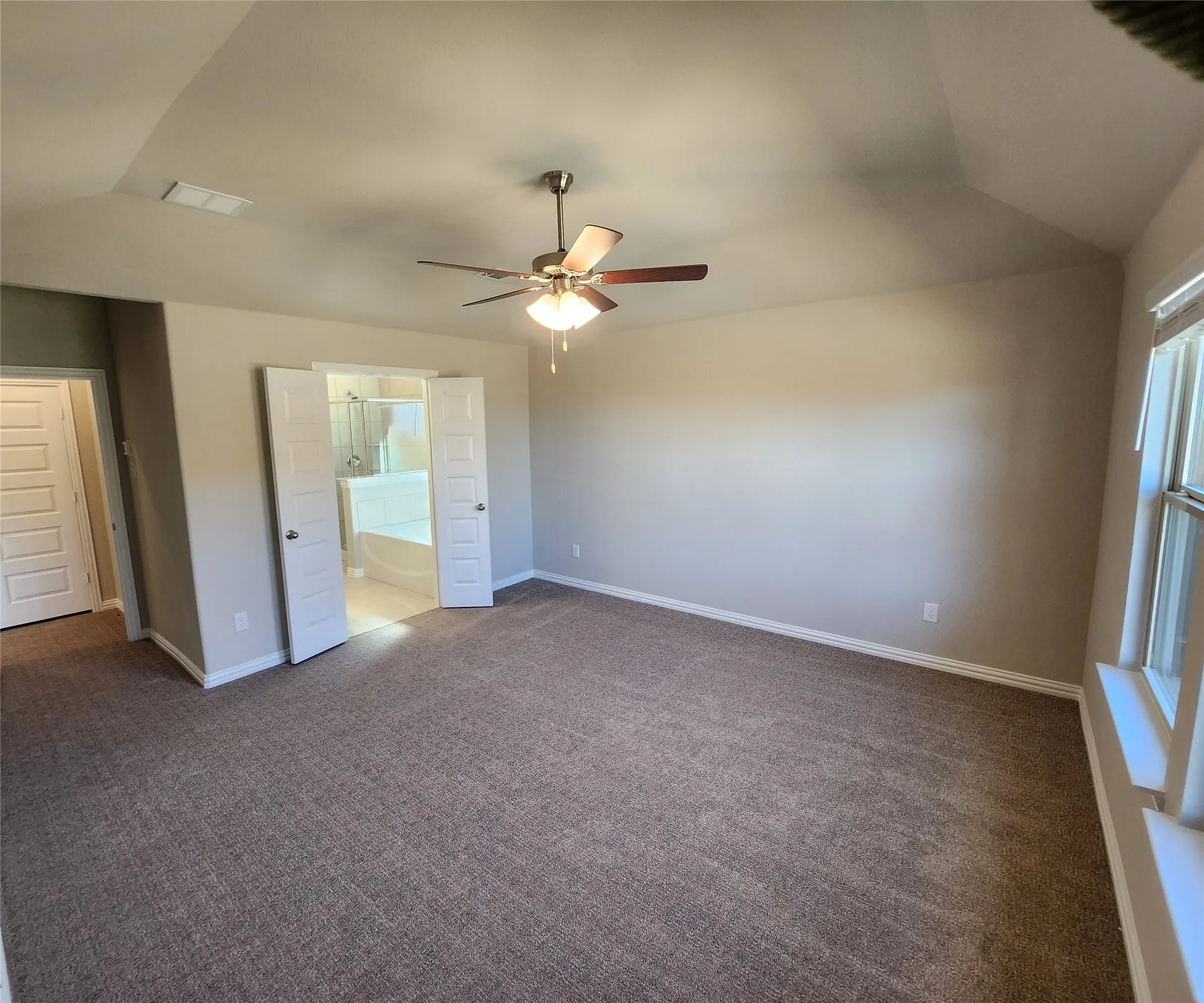 Vaulted ceiling with ceiling fan, neutral carpet, double windows with window sills, and double doors opening to a bathroom with a soaking tub and glass-enclosed shower