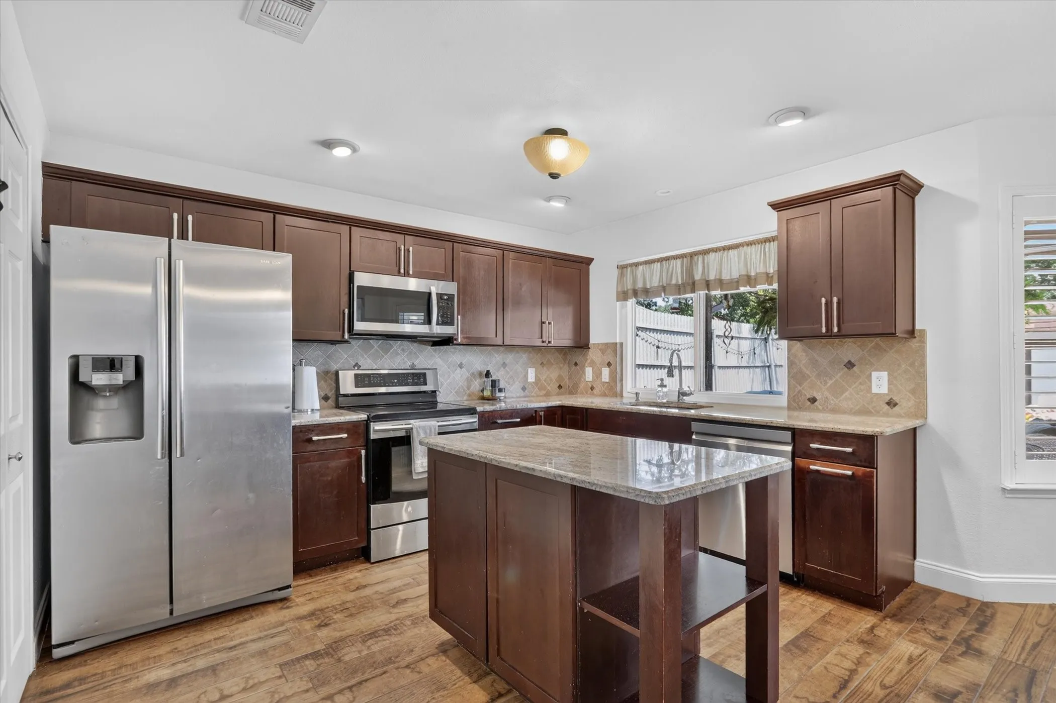 Kitchen featuring wood-finish tile flooring, stainless steel appliances, dark wood cabinetry, light-toned countertops, and a central island
