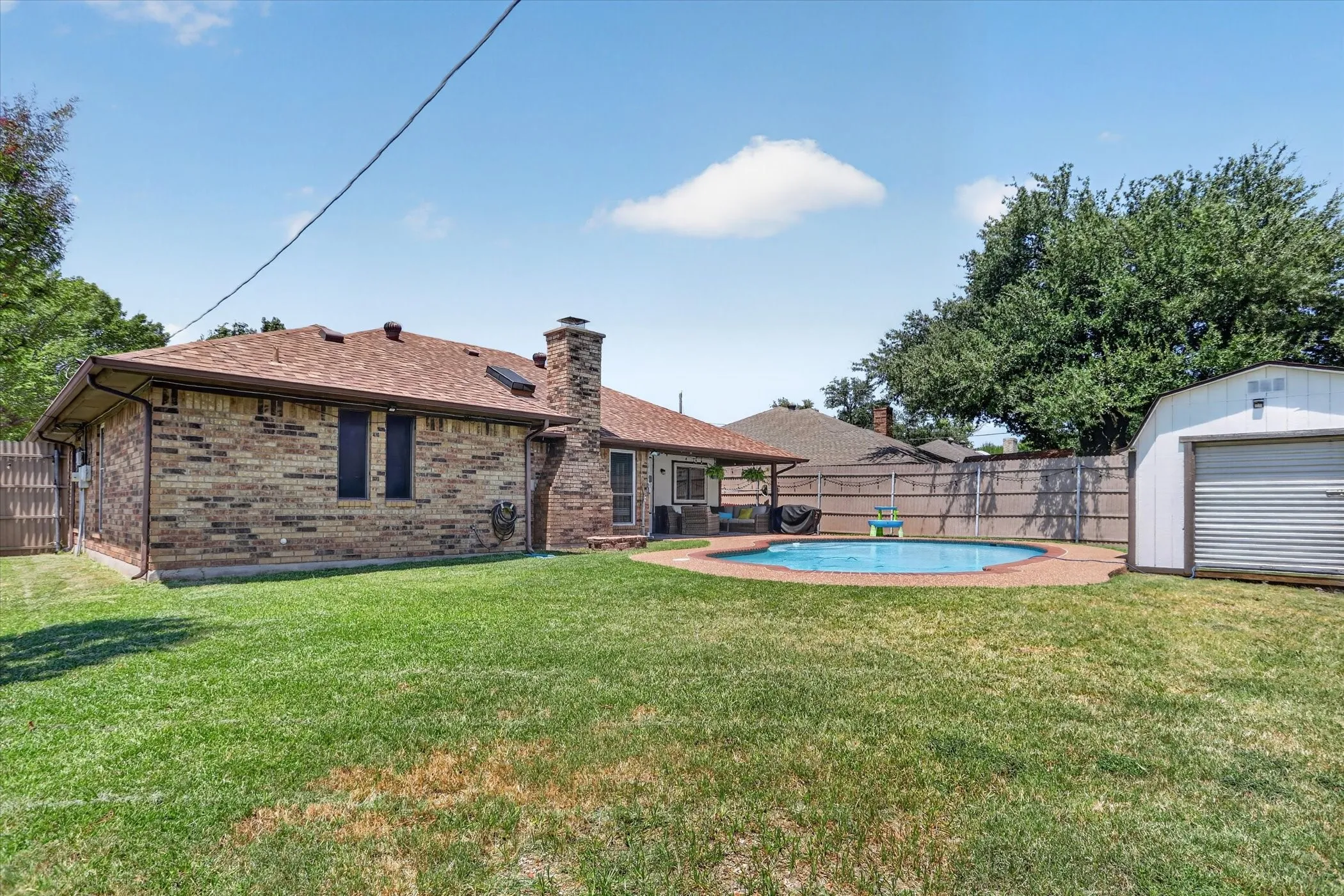 Brick exterior with a covered patio, a circular swimming pool with brick coping, a large storage shed, and a privacy fence