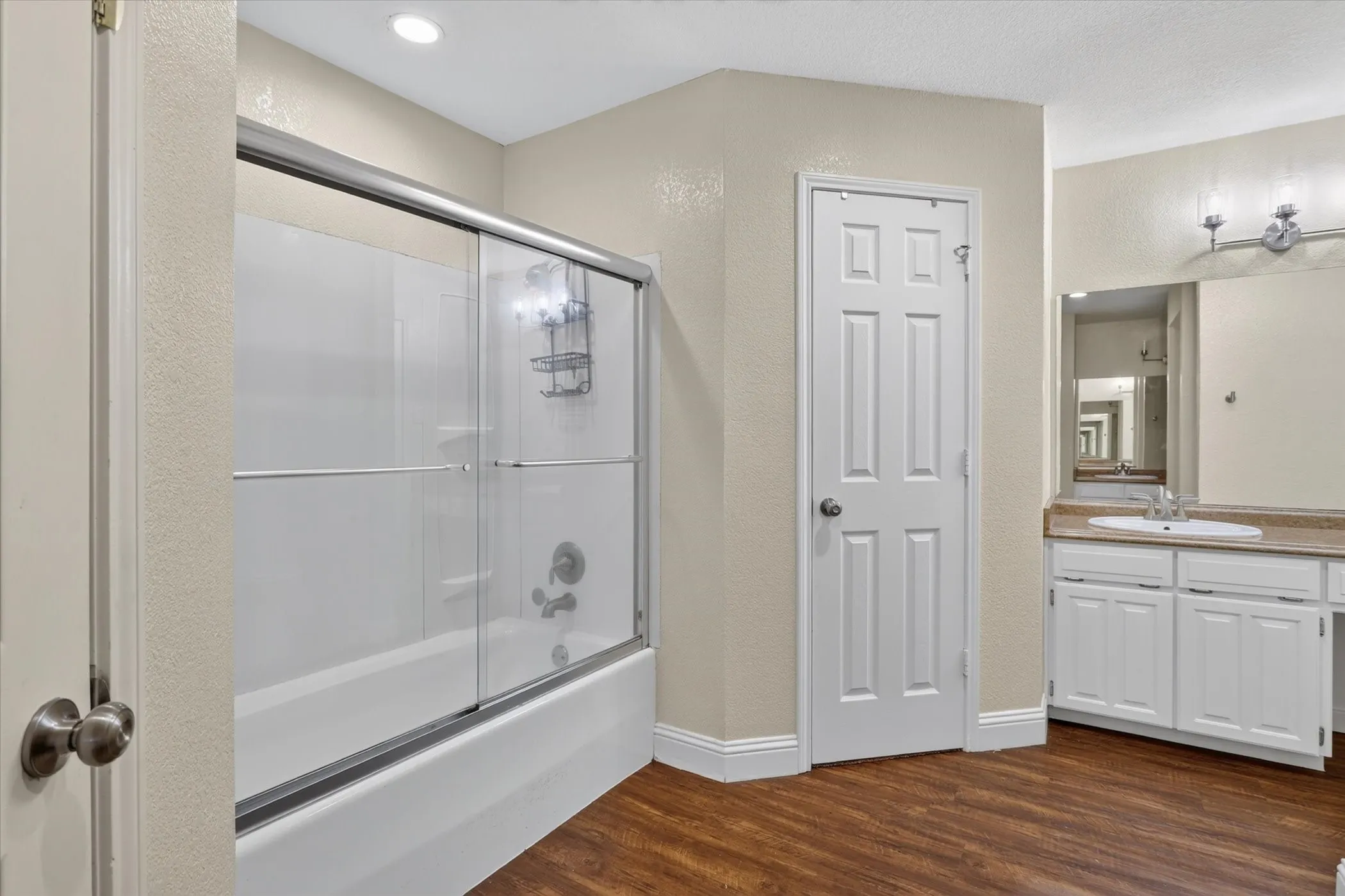Bathtub with sliding glass enclosure, white vanity with integrated sink, stone-finish countertop, and wood-finish flooring