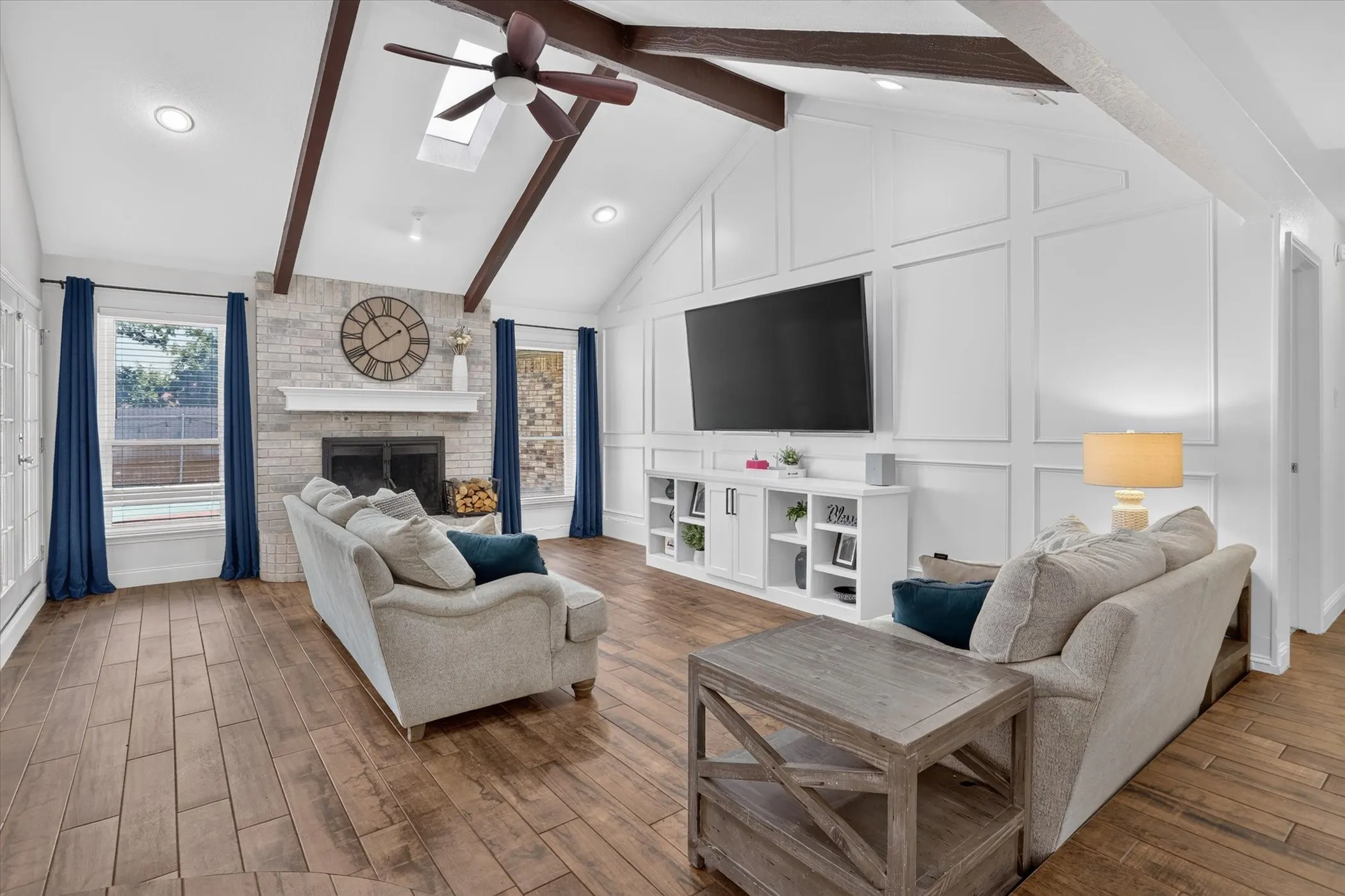 Vaulted ceiling with exposed wood beams and skylight, wood-finish tile flooring, brick-surround fireplace with mantel, built-in media console, and decorative wall paneling
