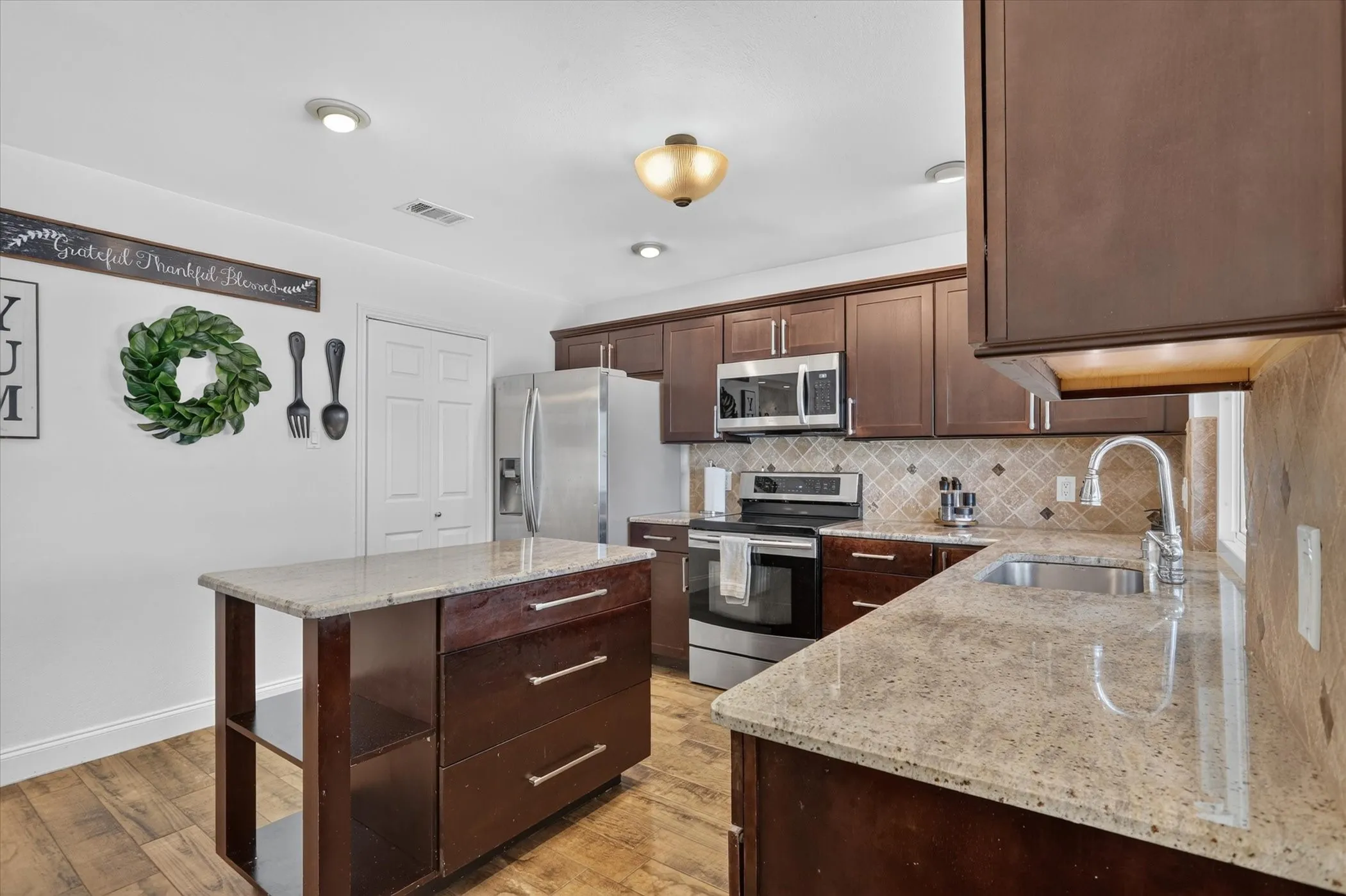 Kitchen featuring wood-finish tile flooring, dark wood cabinetry, light-toned countertops, stainless steel appliances, and an arabesque-patterned backsplash