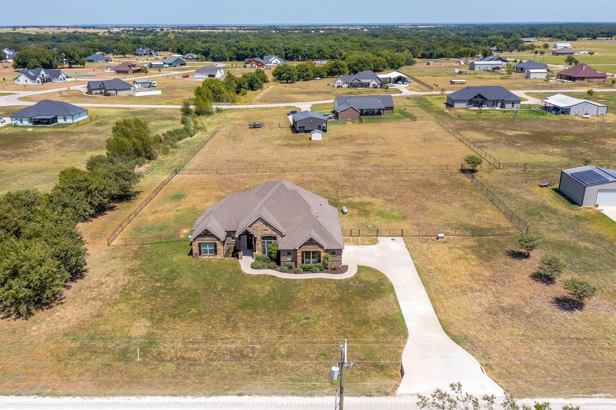 Stucco and stone exterior, concrete driveway, fenced property lines, detached accessory structure, and mature trees on the property perimeter