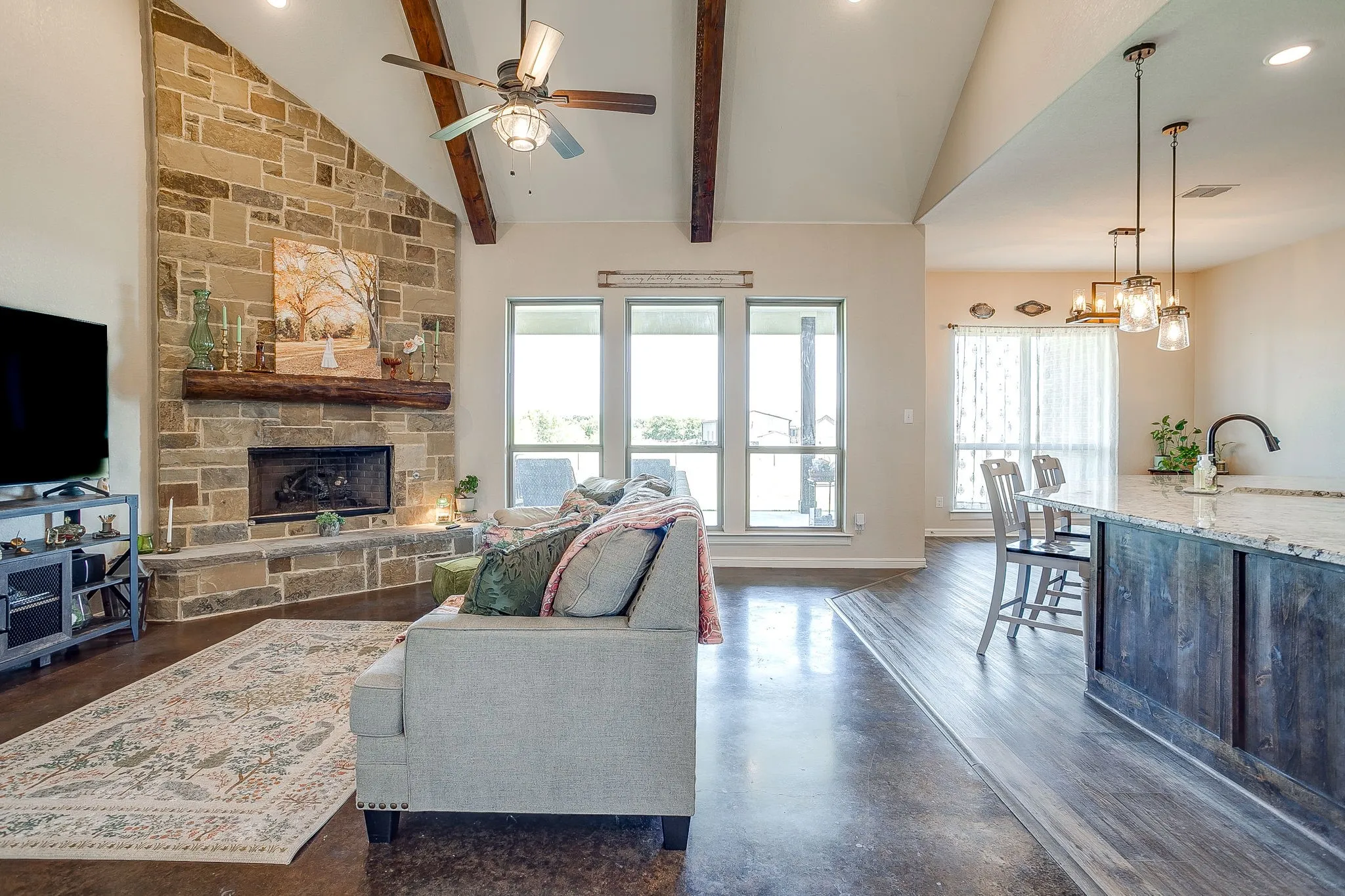 Vaulted ceiling with exposed wood beams, stone-faced fireplace with a wood mantle, large windows providing natural illumination, wood-finish flooring, and a kitchen island with pendant lighting and bar seating