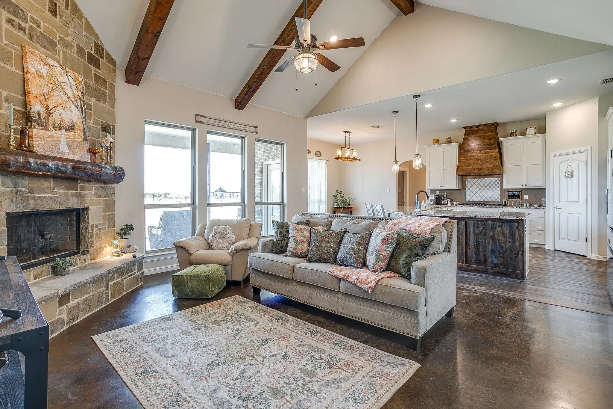 Vaulted ceiling with exposed wood beams, stone fireplace with mantel, polished concrete flooring, white cabinetry, and a kitchen island with stone countertops