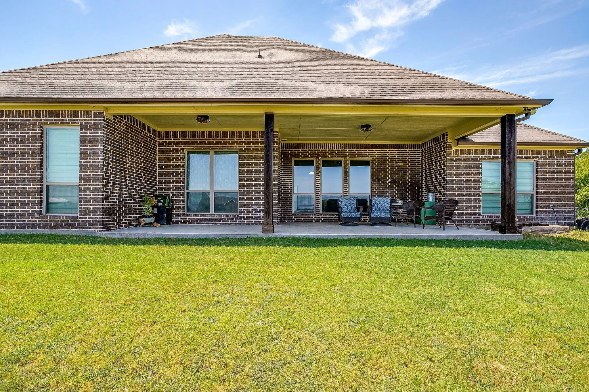 Expansive backyard featuring a manicured lawn, brick exterior, and a covered concrete patio supported by dark wood-tone columns