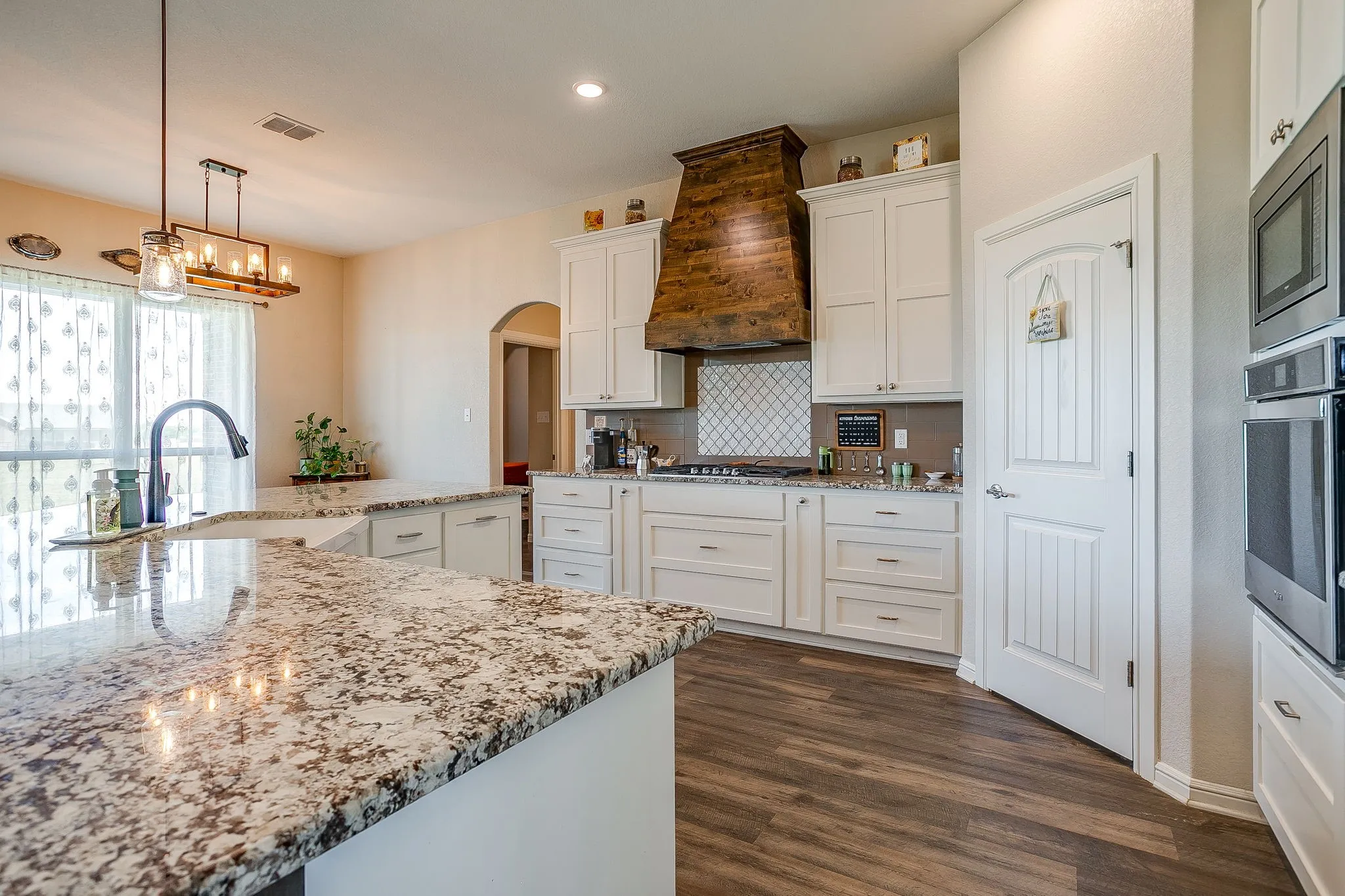 Spacious kitchen featuring an expansive island with a granite countertop, white cabinetry with contemporary pulls, wood-finish flooring, and a decorative wood range hood