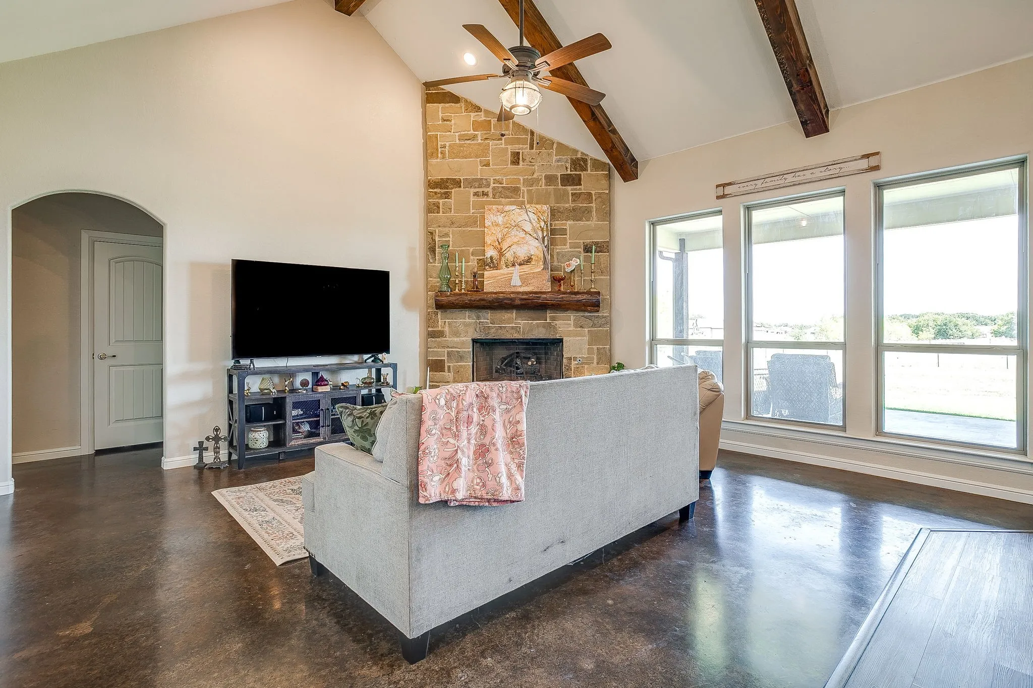 Vaulted ceiling with exposed wood beams, stone-clad fireplace with wood mantel, a ceiling fan with integrated lighting, and multiple large windows