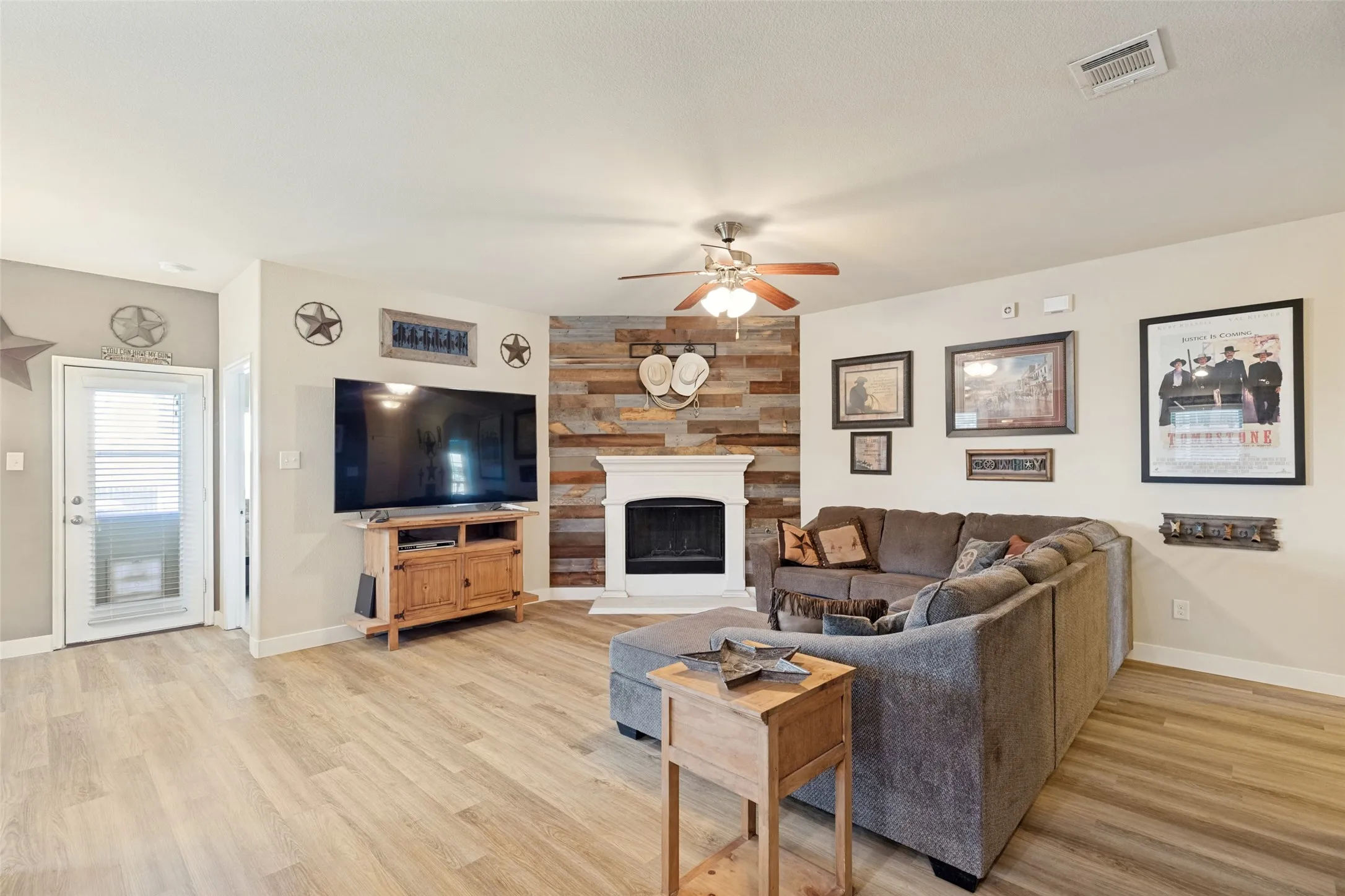Wood-finish flooring throughout, ceiling fan with integrated lighting, white mantel fireplace with wood plank accent wall, recessed lighting.