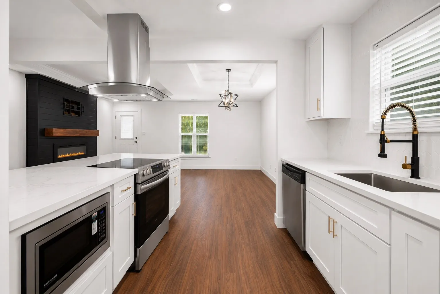 White shaker cabinetry with gold-tone hardware, stainless steel appliances, wood-finish flooring, a contemporary electric fireplace, and a black gooseneck faucet