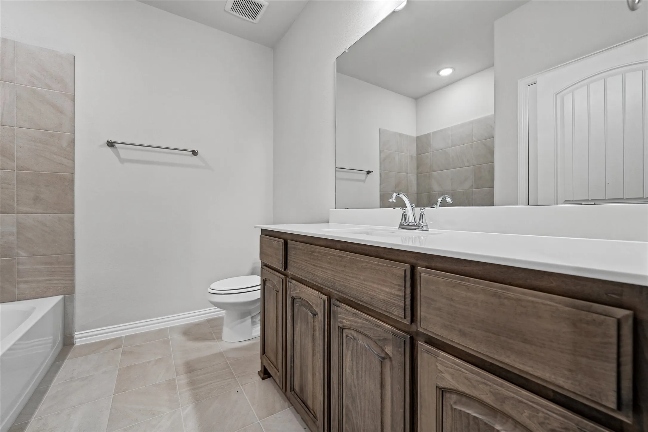 White vanity countertop, wood-finish cabinetry, tile flooring, and a tiled tub surround