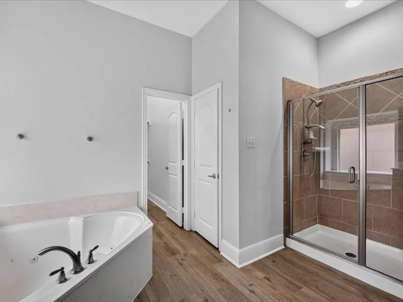 Corner soaking tub with dark bronze fixtures, glass-enclosed shower with tiled surround, wood-finish flooring, and light gray wall paint