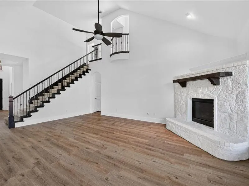 Vaulted ceiling with a ceiling fan, wood-finish flooring, stone fireplace with a wood mantle, and a staircase with wrought iron balusters