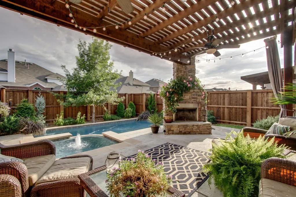 Wood pergola with string lights, stone fireplace, swimming pool with water feature, and a privacy fence