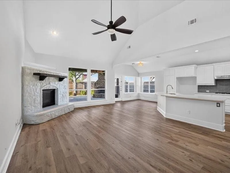 Vaulted ceilings, wood-finish flooring, stone fireplace, white kitchen cabinetry, and large windows