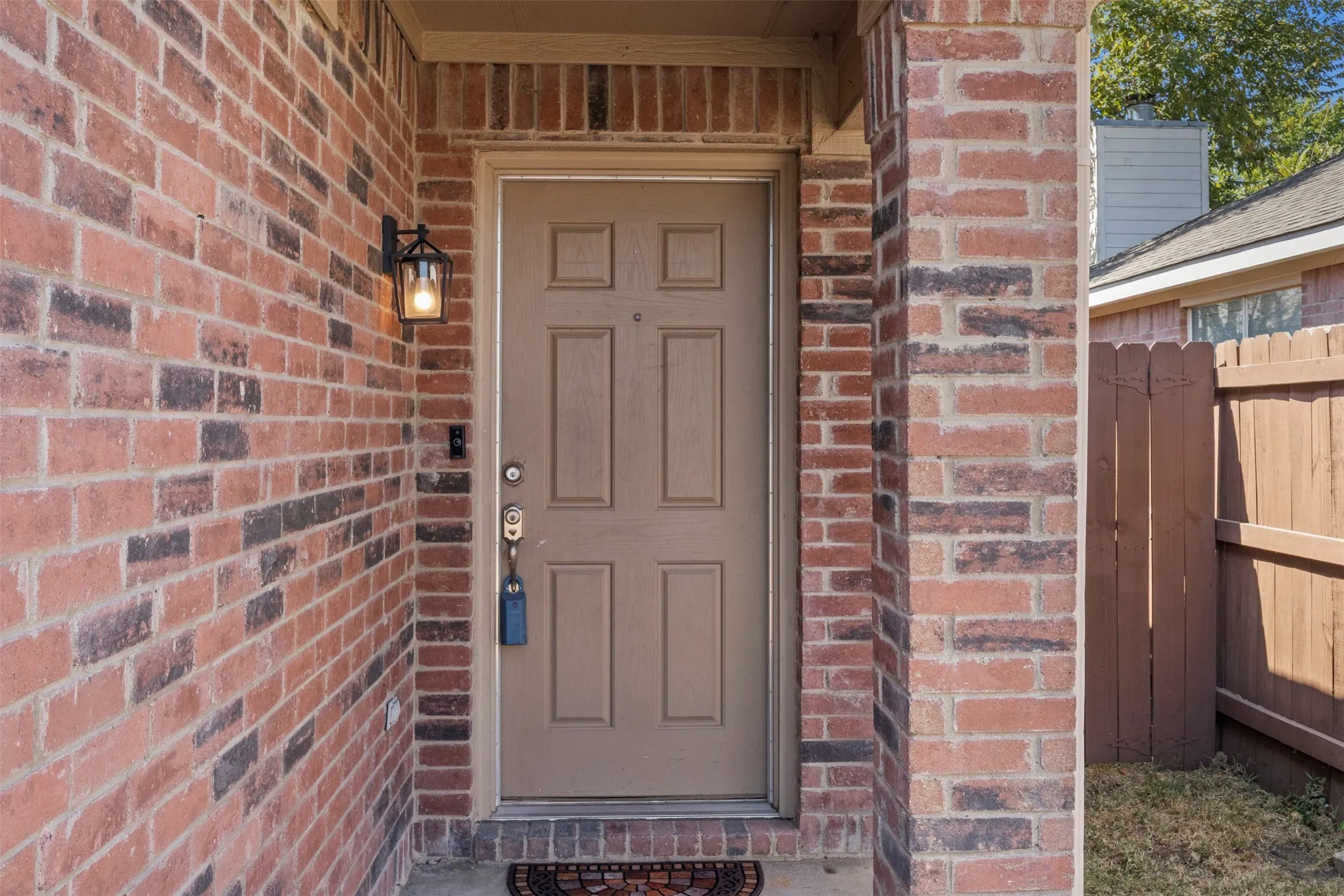 Brick exterior facade with a paneled entry door, exterior wall lantern, and partial wood privacy fence