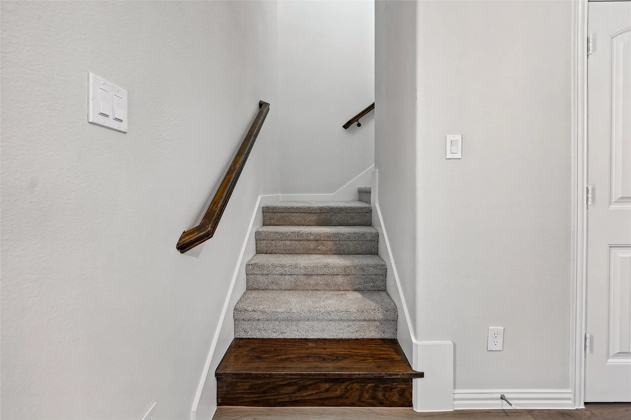 Staircase featuring wood-finish treads, carpeted risers, white painted walls, and a wood-finish handrail with metal brackets
