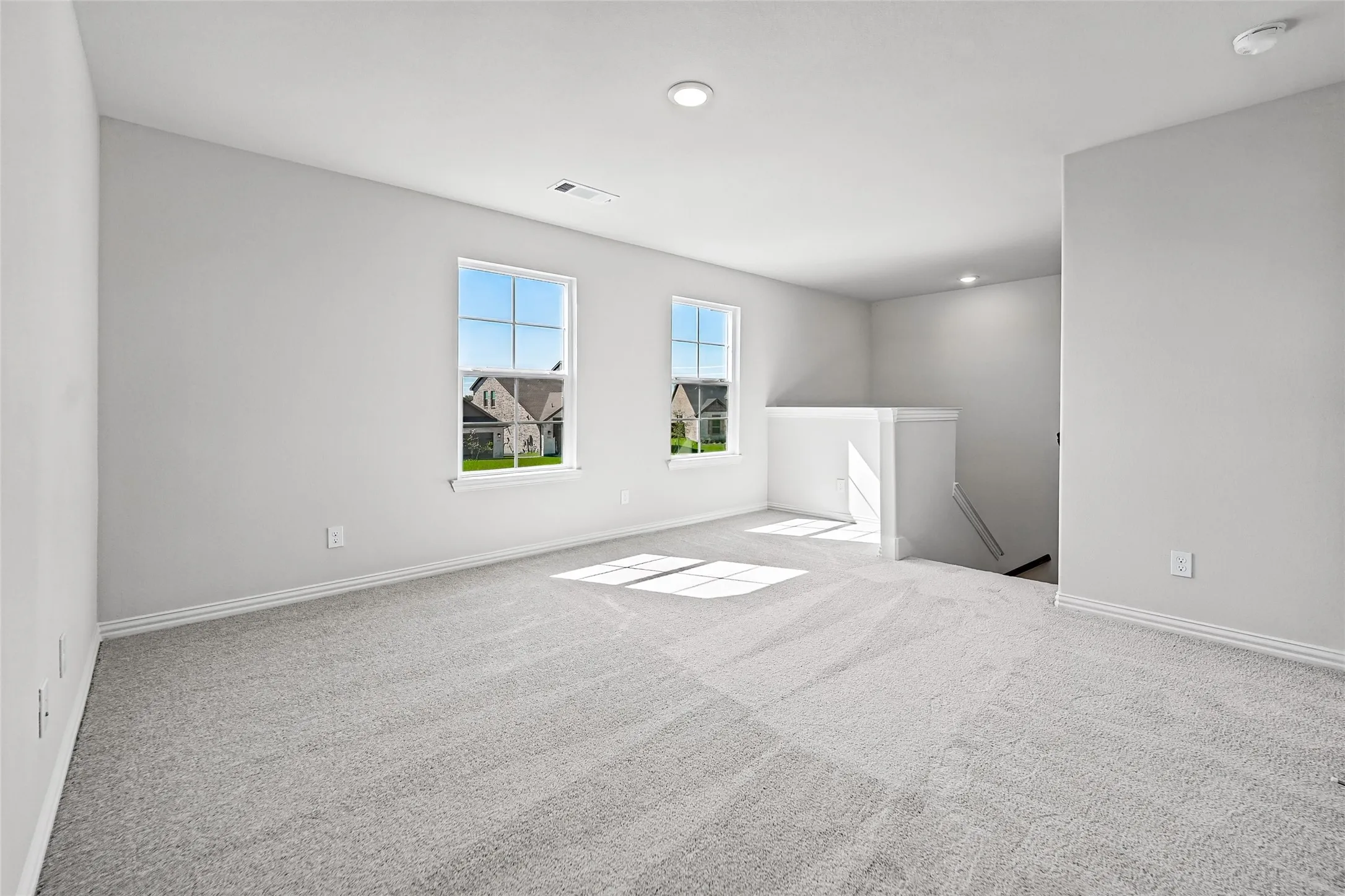 Expansive room featuring light gray carpet flooring, two large windows, recessed lighting, and a white stair railing