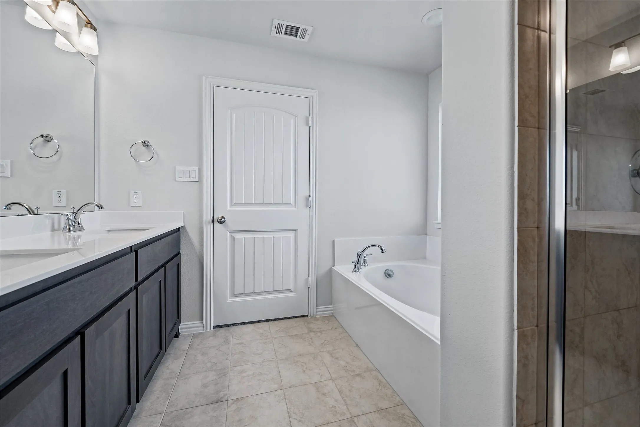 Dual vanity with white countertops, wood-finish cabinetry, tile flooring, built-in bathtub, and a glass-enclosed shower