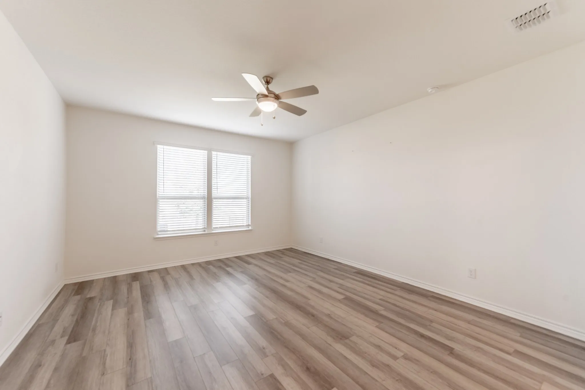 Spacious room featuring light wood-finish flooring, a ceiling fan with integrated lighting, and a large window with blinds