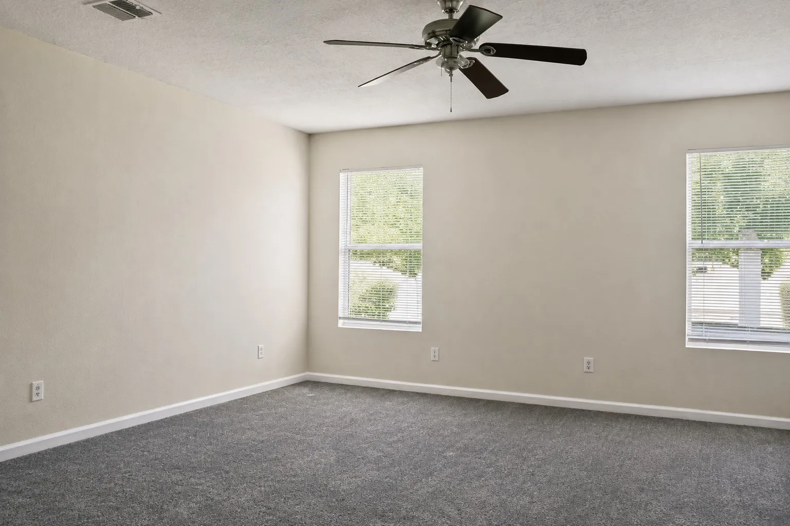 Neutral painted walls with a ceiling fan, two windows, and gray carpet flooring
