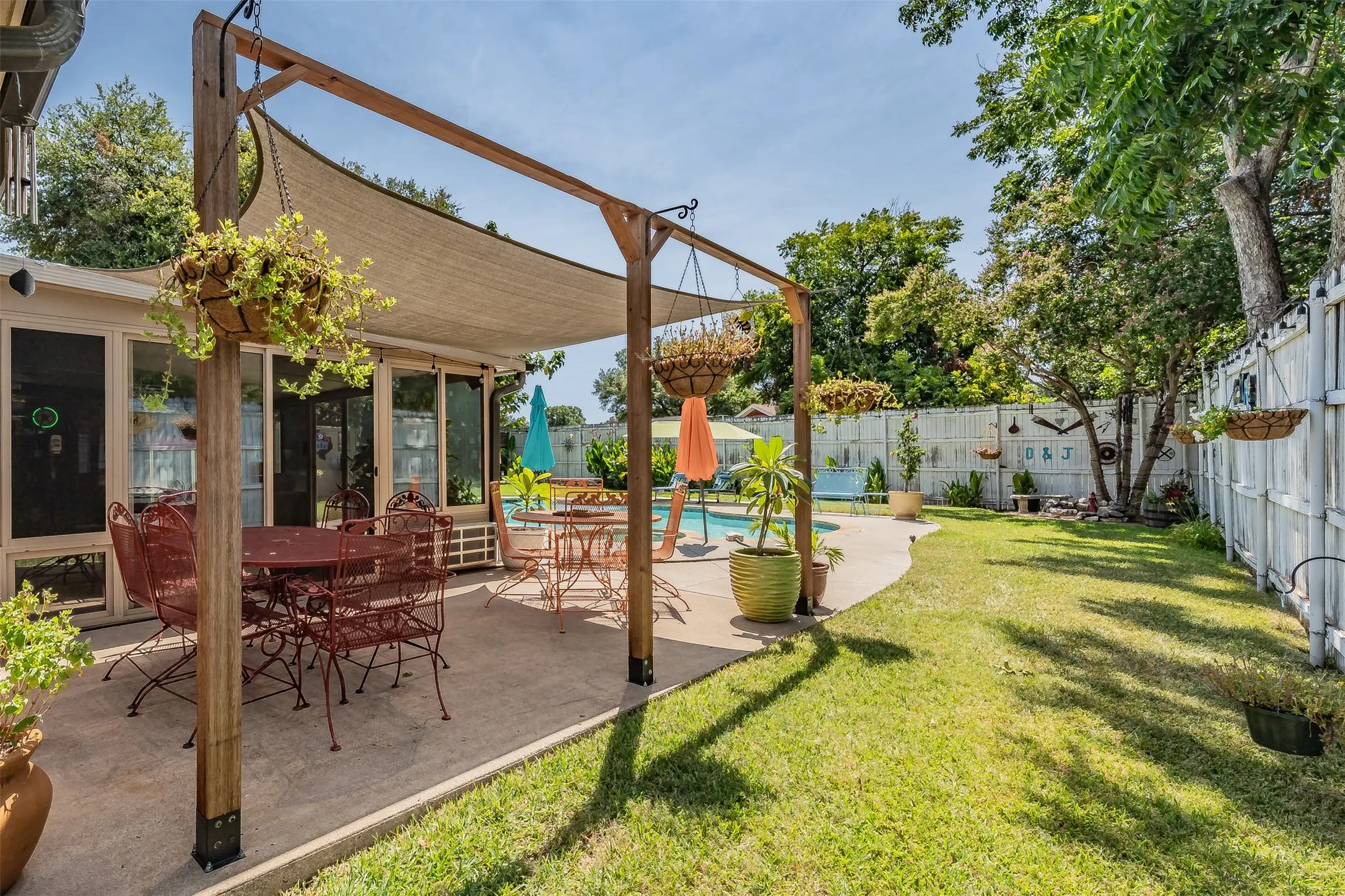 Expansive concrete patio with a wooden pergola and shade sail, bordered by a manicured lawn