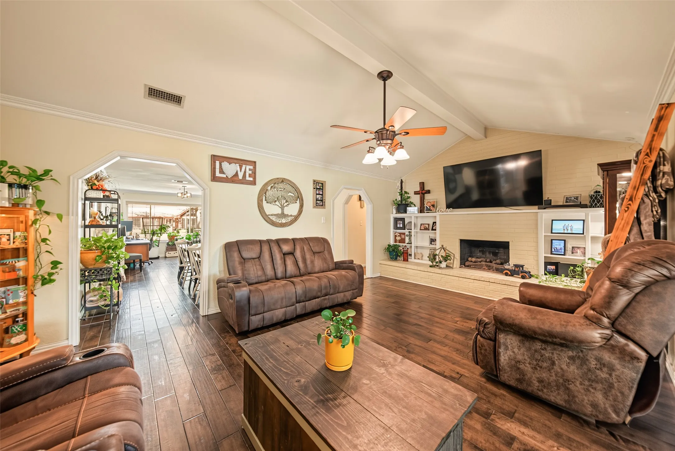 Vaulted ceiling with exposed beams, natural wood flooring, built-in shelving, fireplace with brick surround, and a ceiling fan