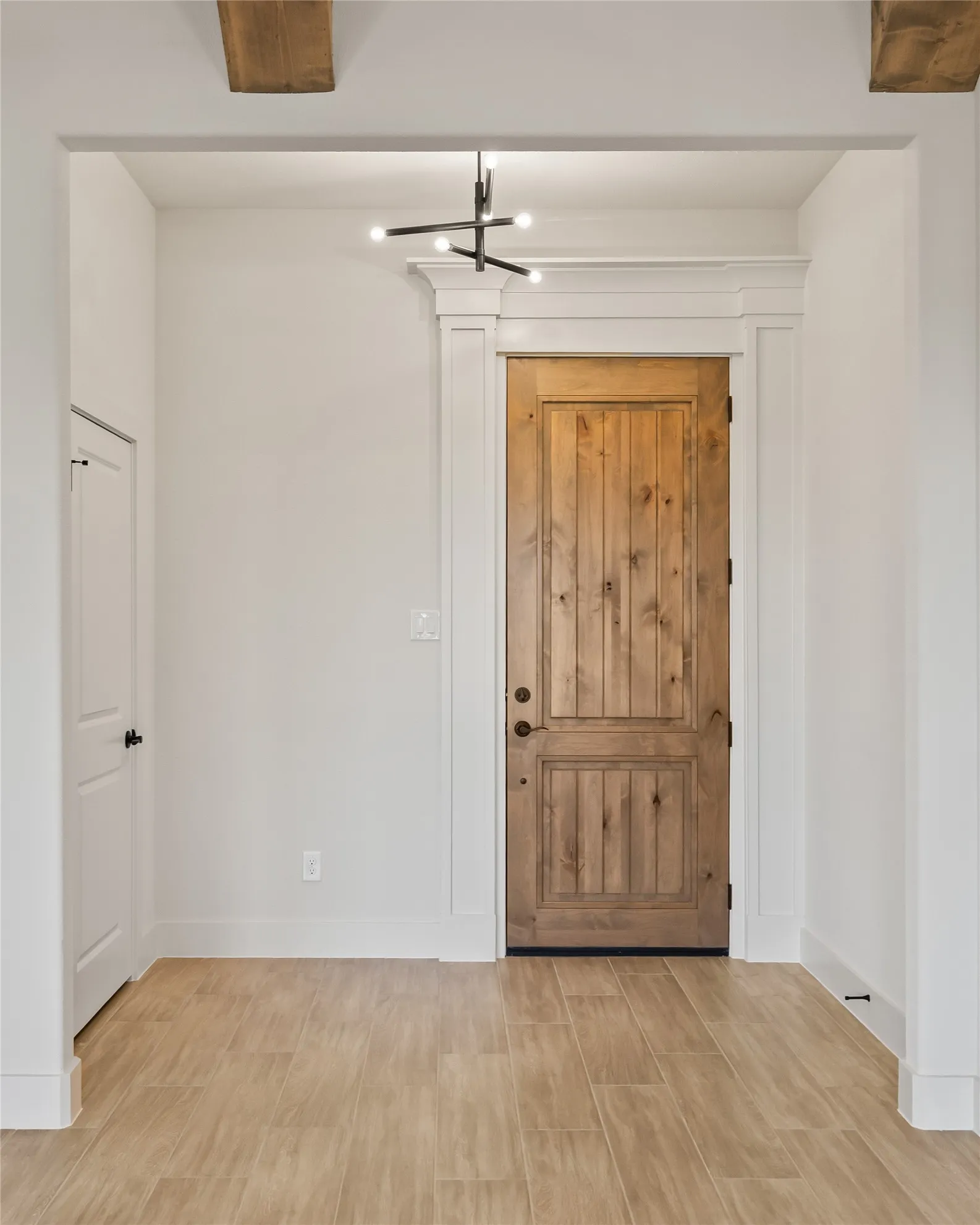 Warm wood-finish flooring, natural wood door with dark hardware, white trim, exposed wood ceiling beams, and a modern ceiling light fixture