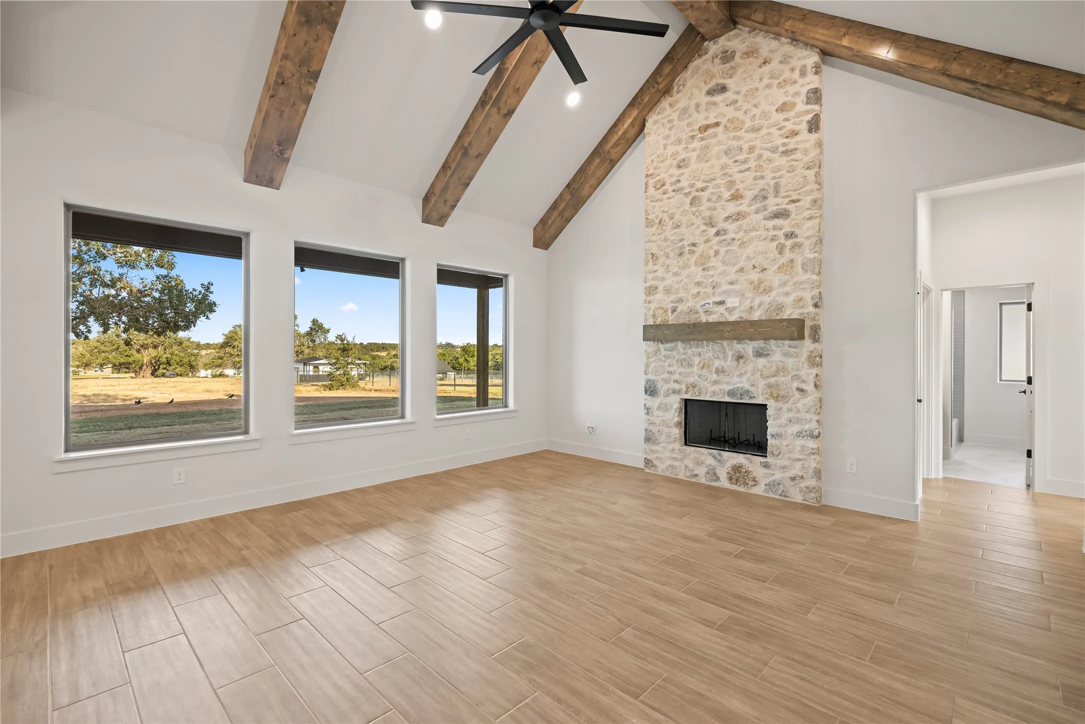 Vaulted ceiling with exposed wood beams, stone-faced fireplace with wood mantel, multiple large windows, wood-finish flooring, and a modern ceiling fan