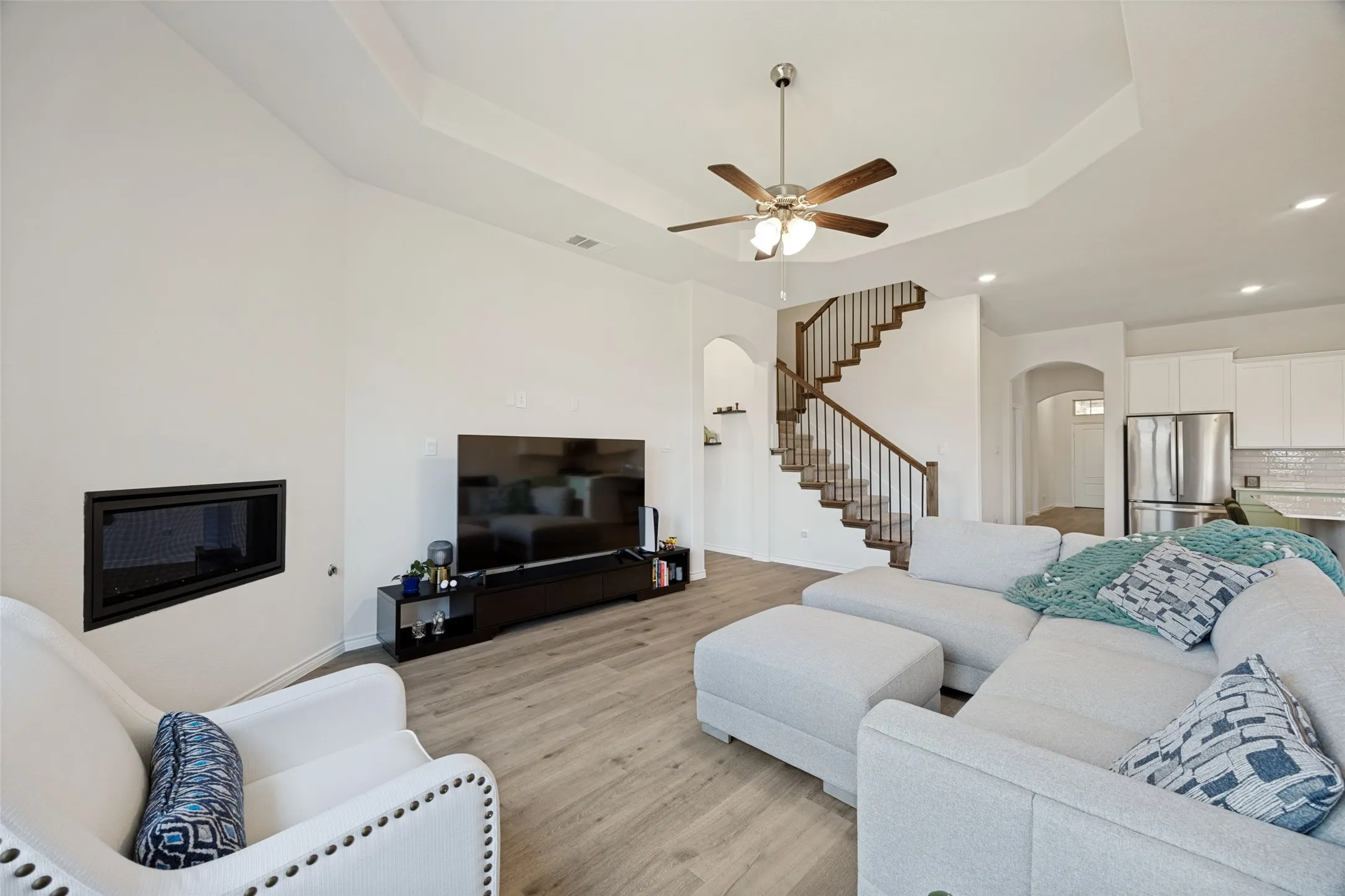 Recessed fireplace, wood-finish flooring, tray ceiling, ceiling fan, and staircase with wood handrails