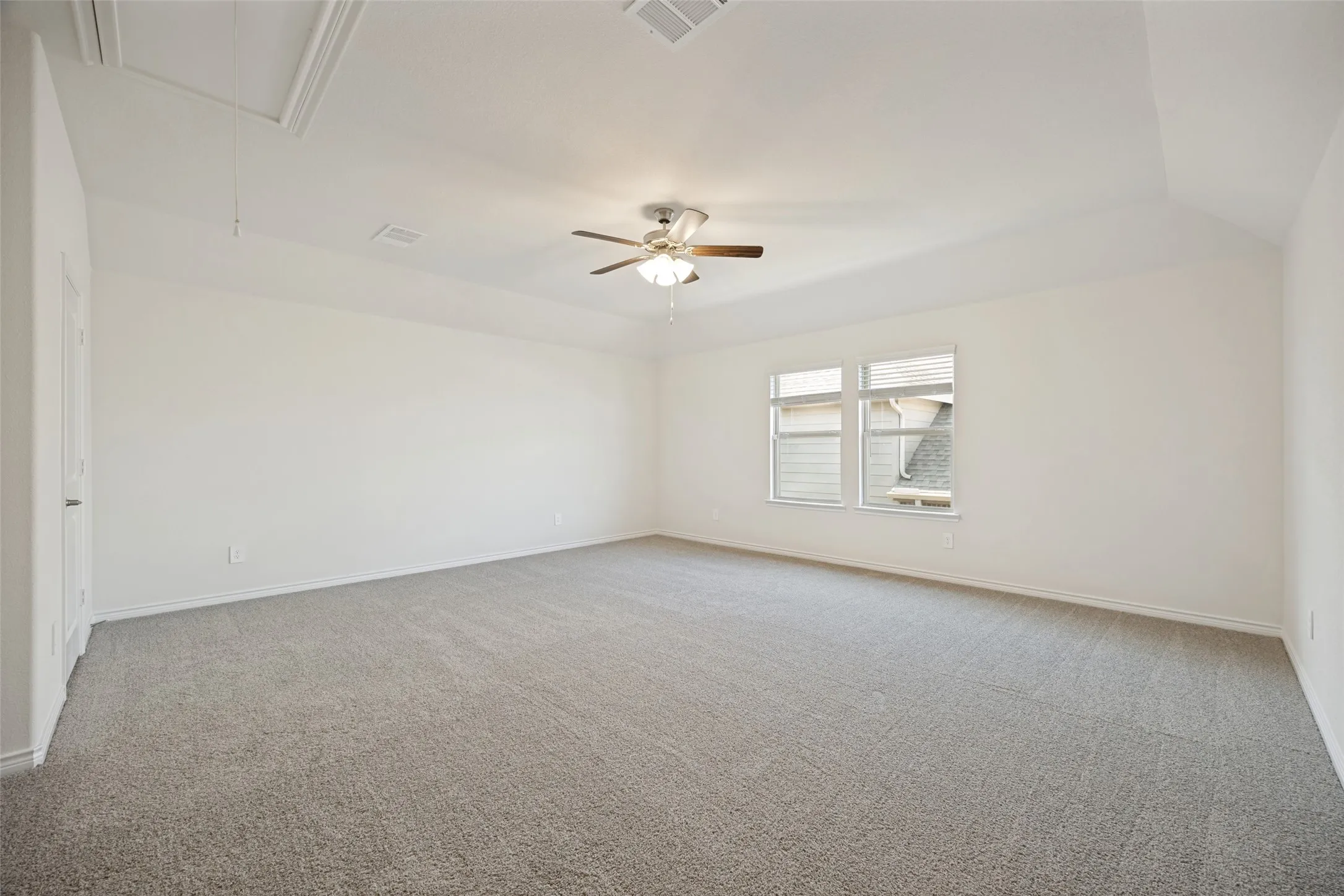 Vaulted ceiling with an attic access panel, two windows with blinds, a ceiling fan with integrated lighting, and neutral-toned carpeting