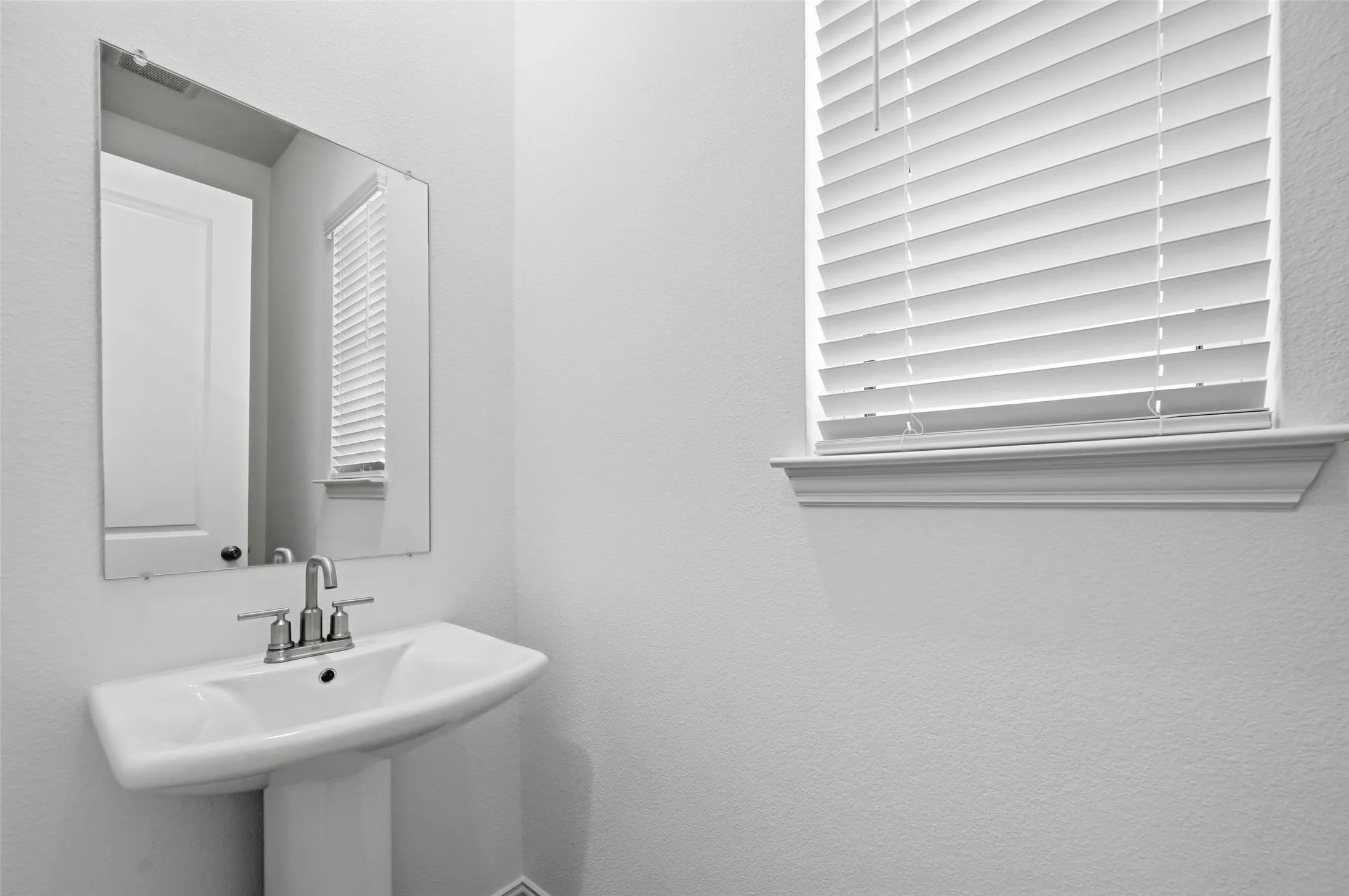 Pedestal sink with a polished chrome faucet, rectangular mirror, and a window featuring white horizontal blinds