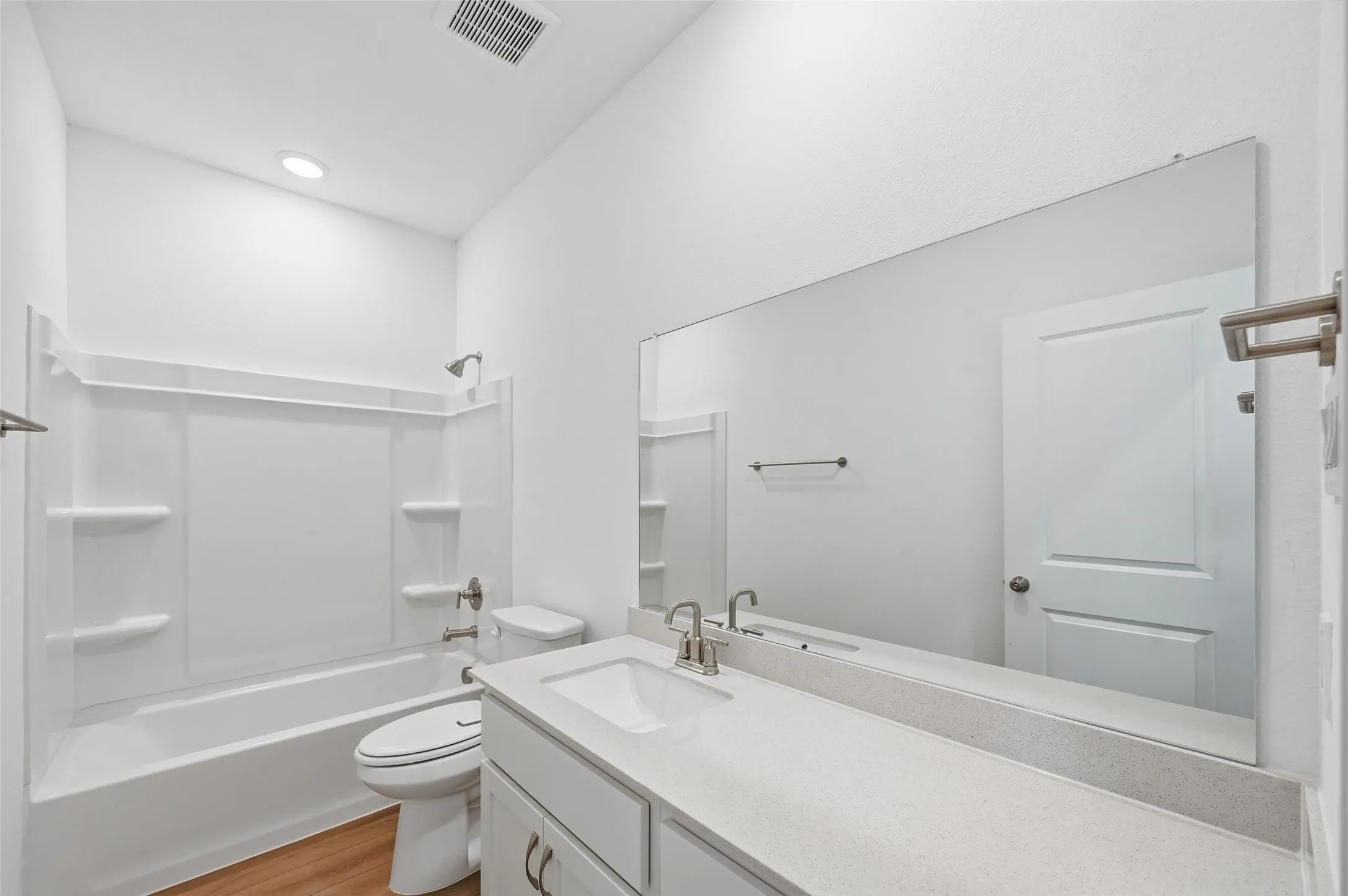 White vanity with a single sink and brushed nickel faucet, large wall-mounted mirror, white bathtub with a shower surround, and wood-finish flooring