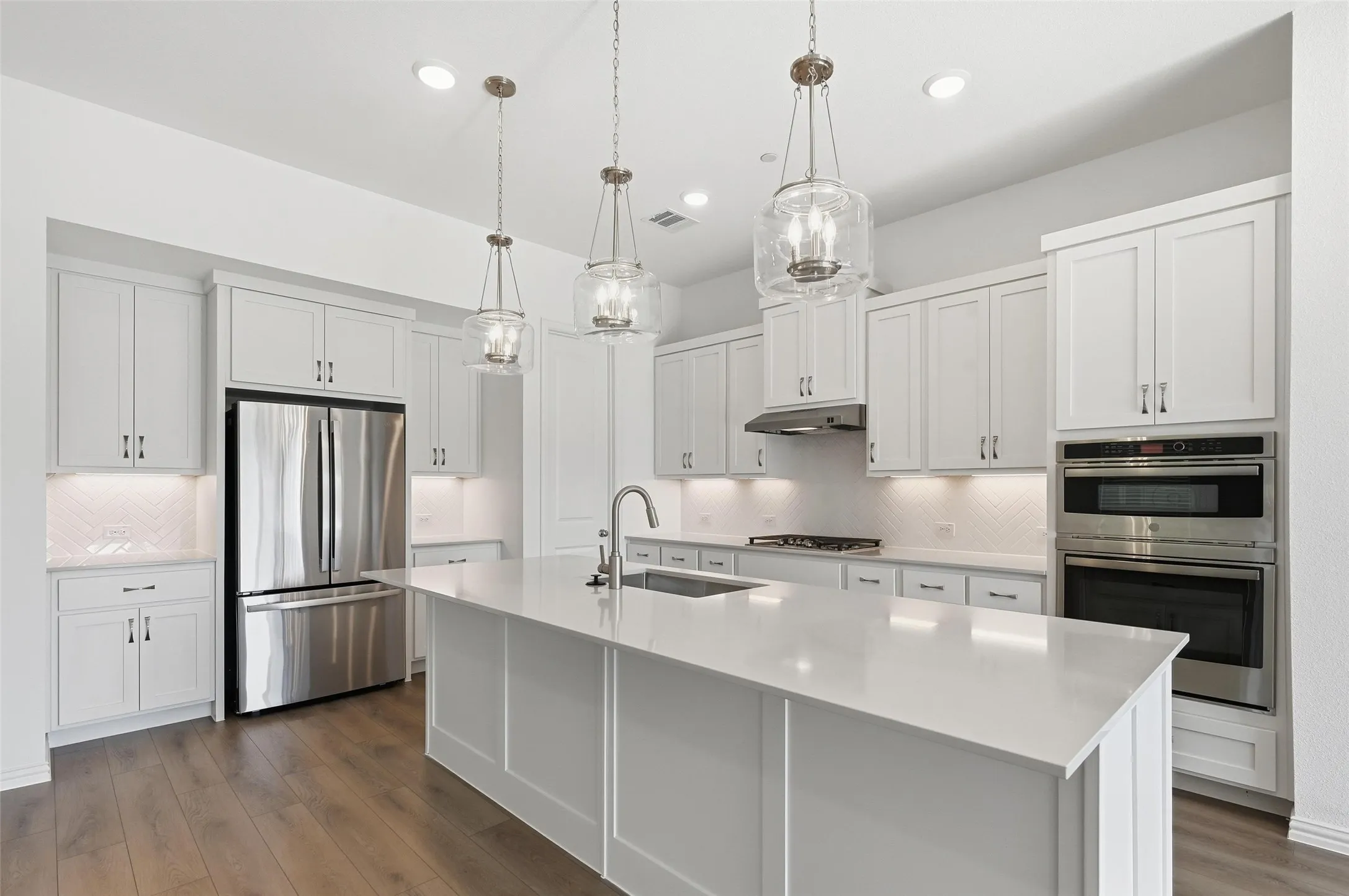 Kitchen featuring white shaker cabinetry, stainless steel appliances, a large center island with a sink, pendant lighting, and wood-finish flooring