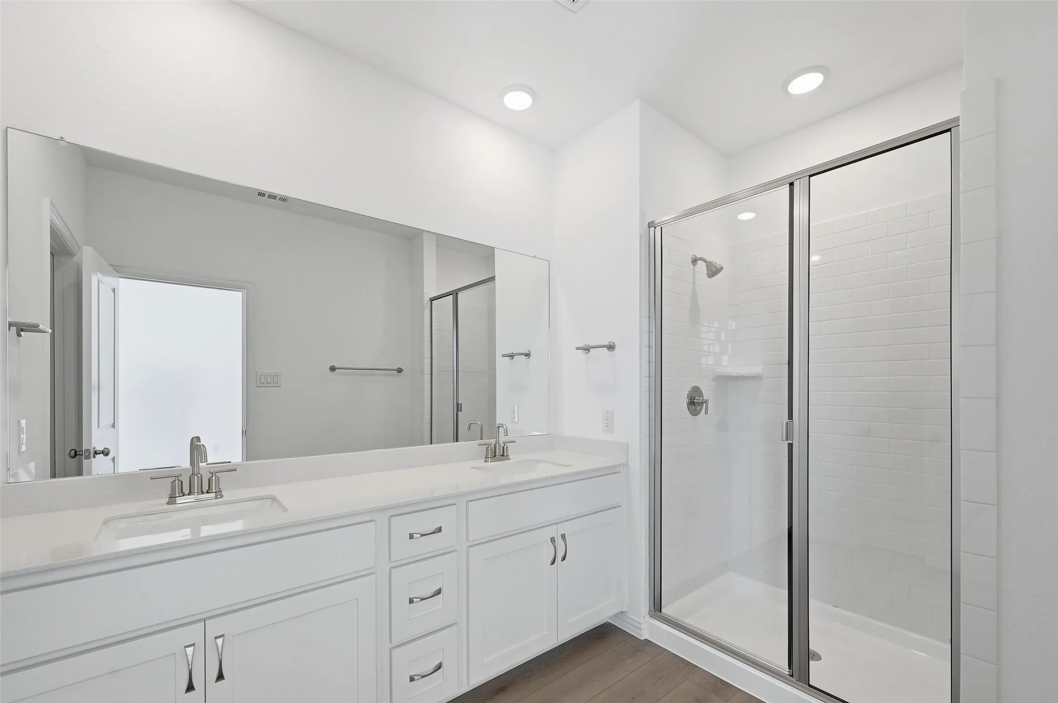 Dual vanity with undermount sinks and chrome fixtures, expansive mirror, wood-finish flooring, and a glass-enclosed shower with subway tile