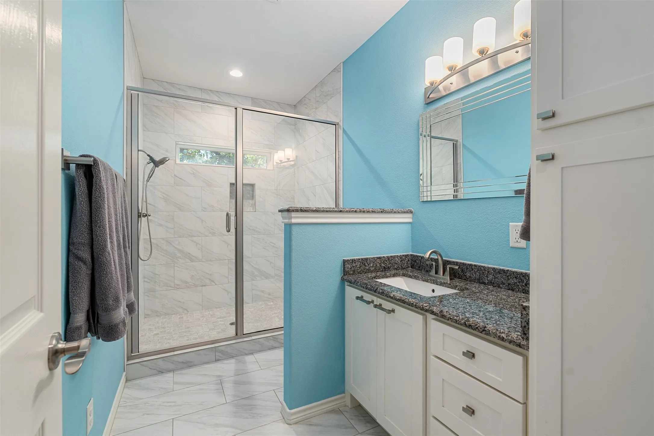 Glass-enclosed shower with a handheld shower fixture, white vanity with a granite-style countertop, and a built-in storage cabinet