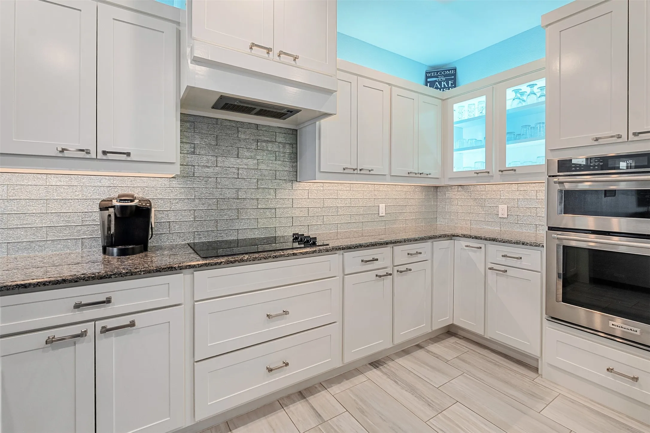 White shaker cabinetry, dark speckled countertops, tile backsplash, built-in stainless steel double ovens, and tile flooring
