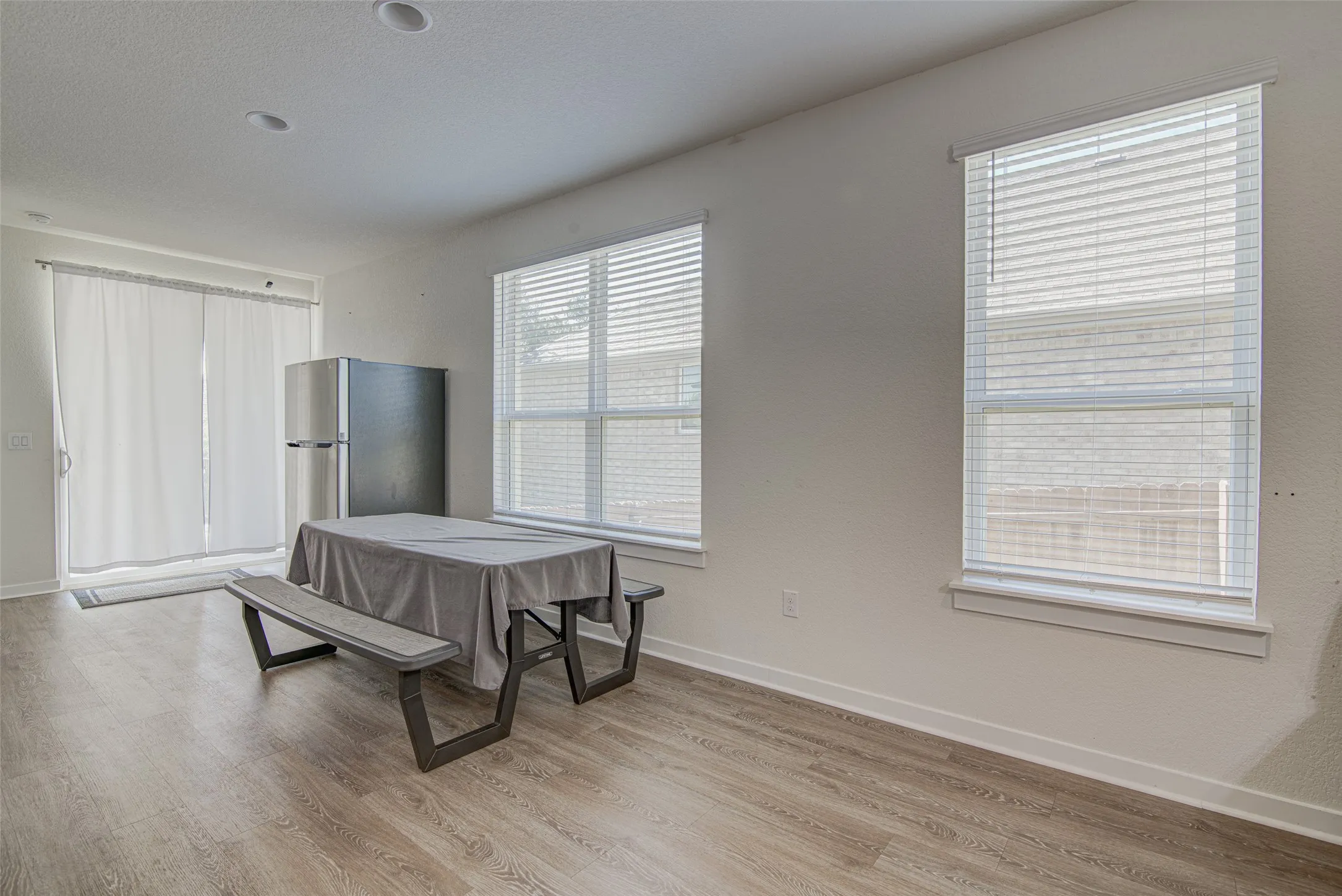 Recessed ceiling lighting, wood-finish flooring, sliding glass door, multiple windows with blinds, and a stainless steel refrigerator