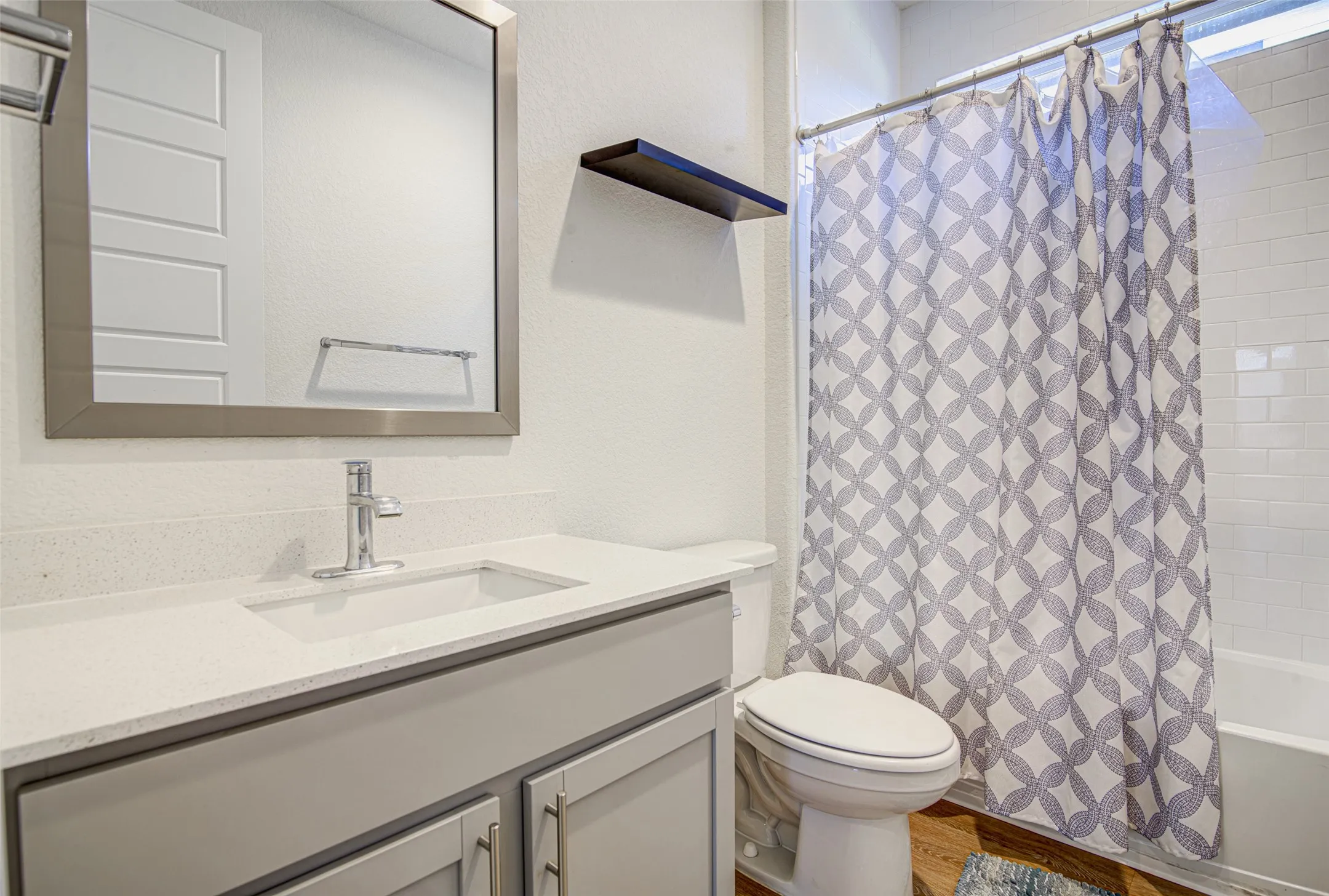 Bathroom featuring a light-colored vanity with a built-in sink, a framed mirror, a toilet, a shower with white subway tile surround, and wood-finish flooring