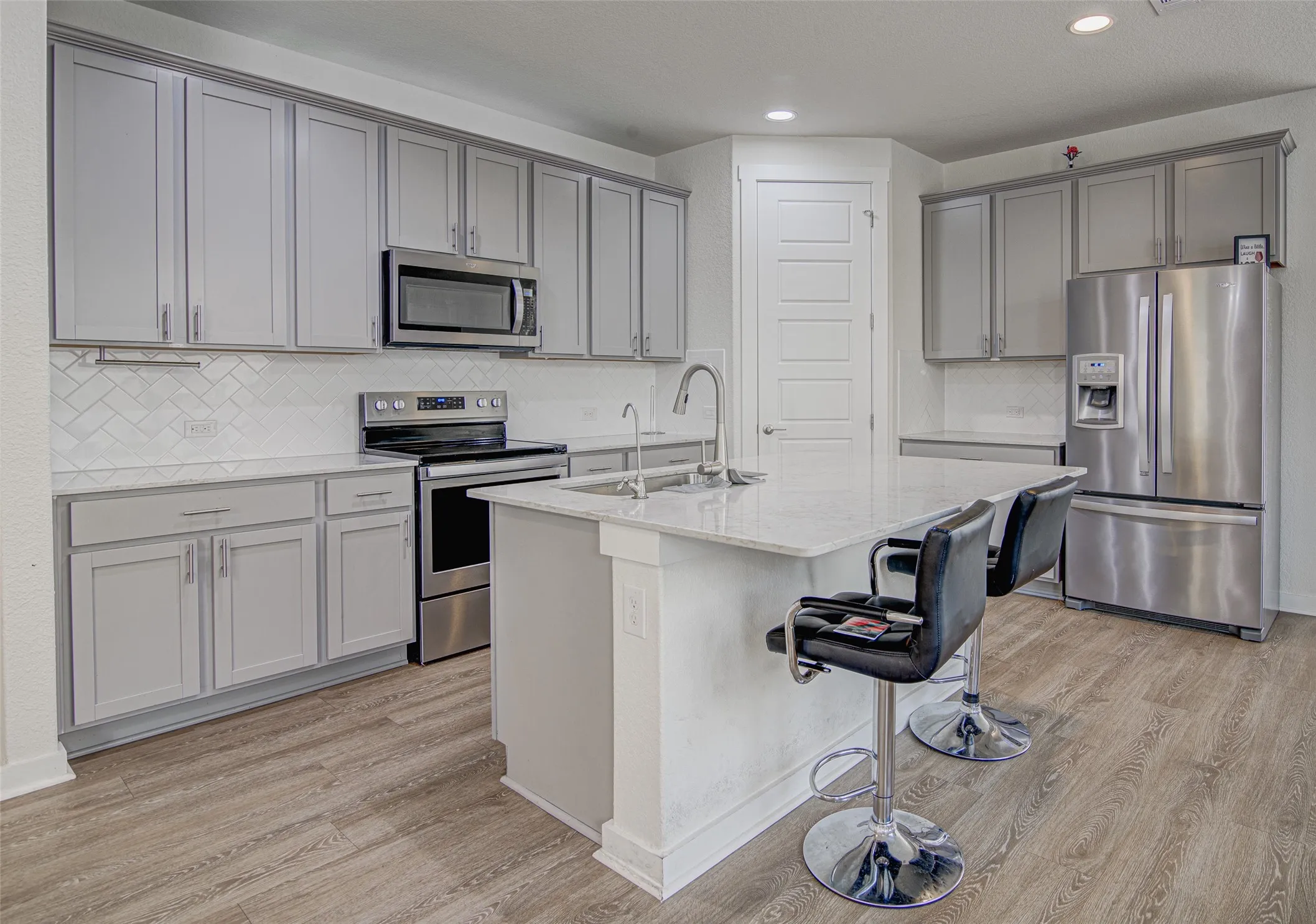 Kitchen with light grey cabinetry, stainless steel appliances, a center island with sink, quartz countertops, and wood-finish flooring