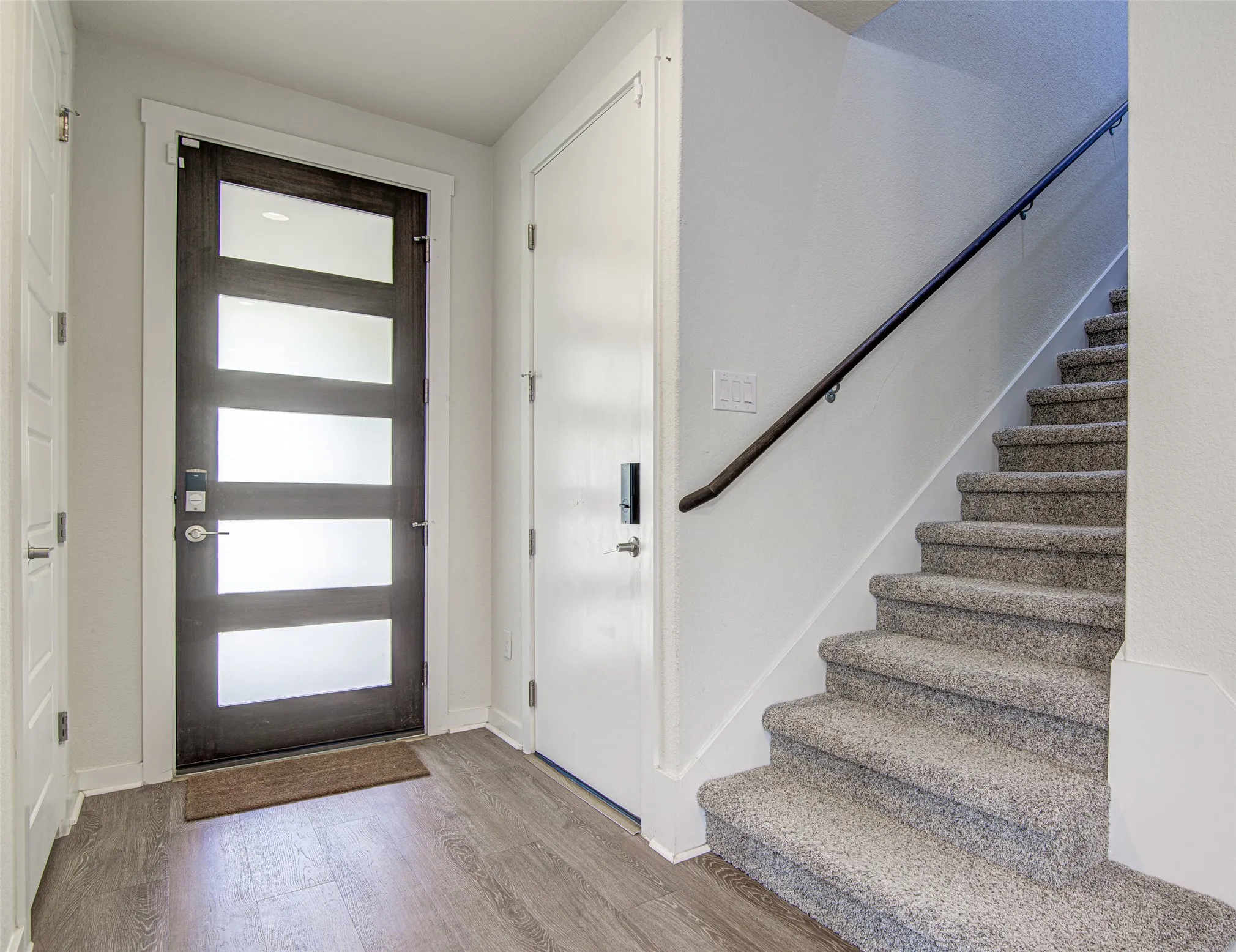 Entryway featuring wood-finish flooring, a contemporary dark-framed door with frosted glass panels, and a carpeted staircase with a dark handrail