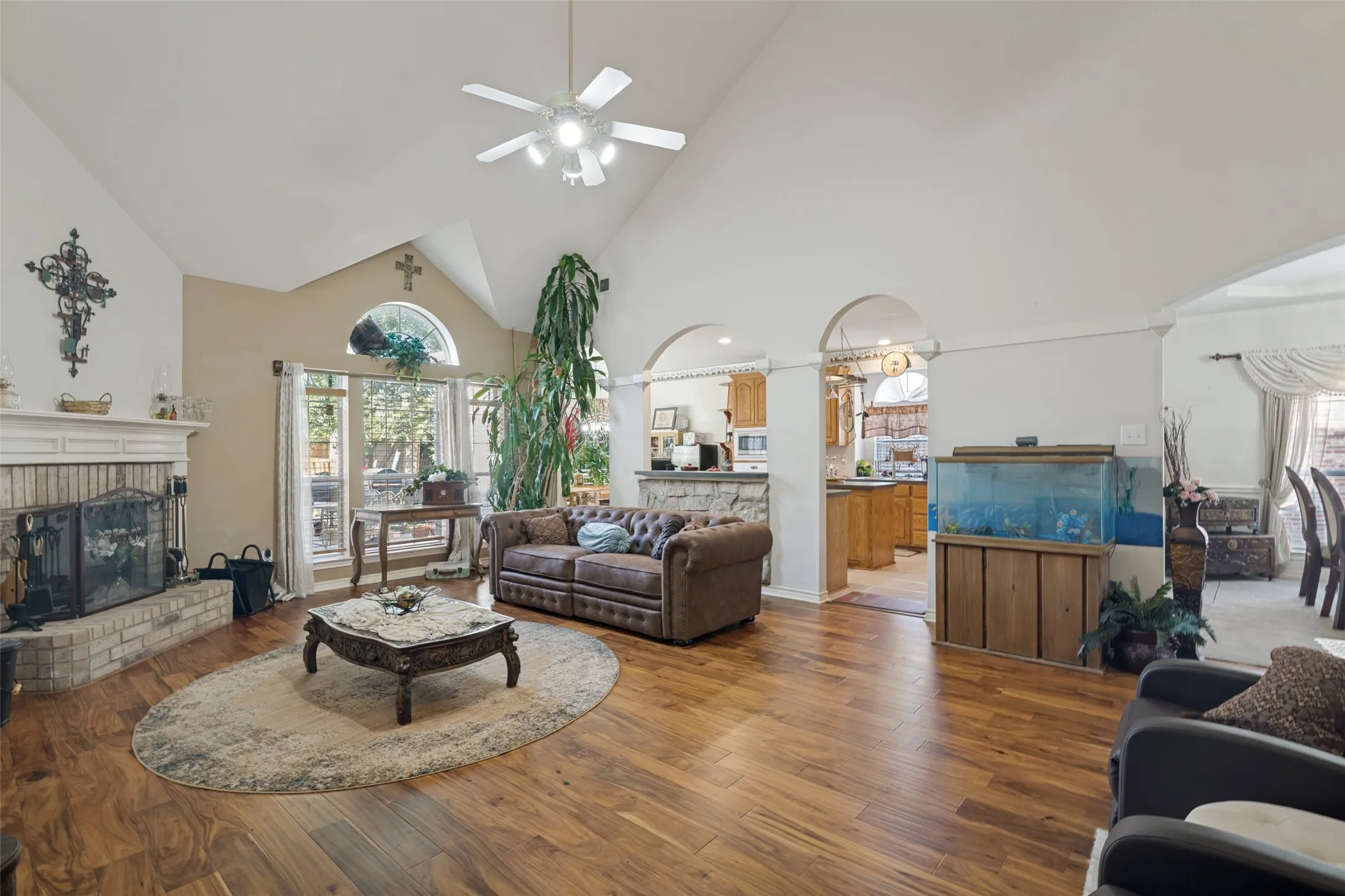 Vaulted ceiling with a ceiling fan, wood-finish flooring, brick-surround fireplace with a mantel, large windows with an arched transom, and arched doorways