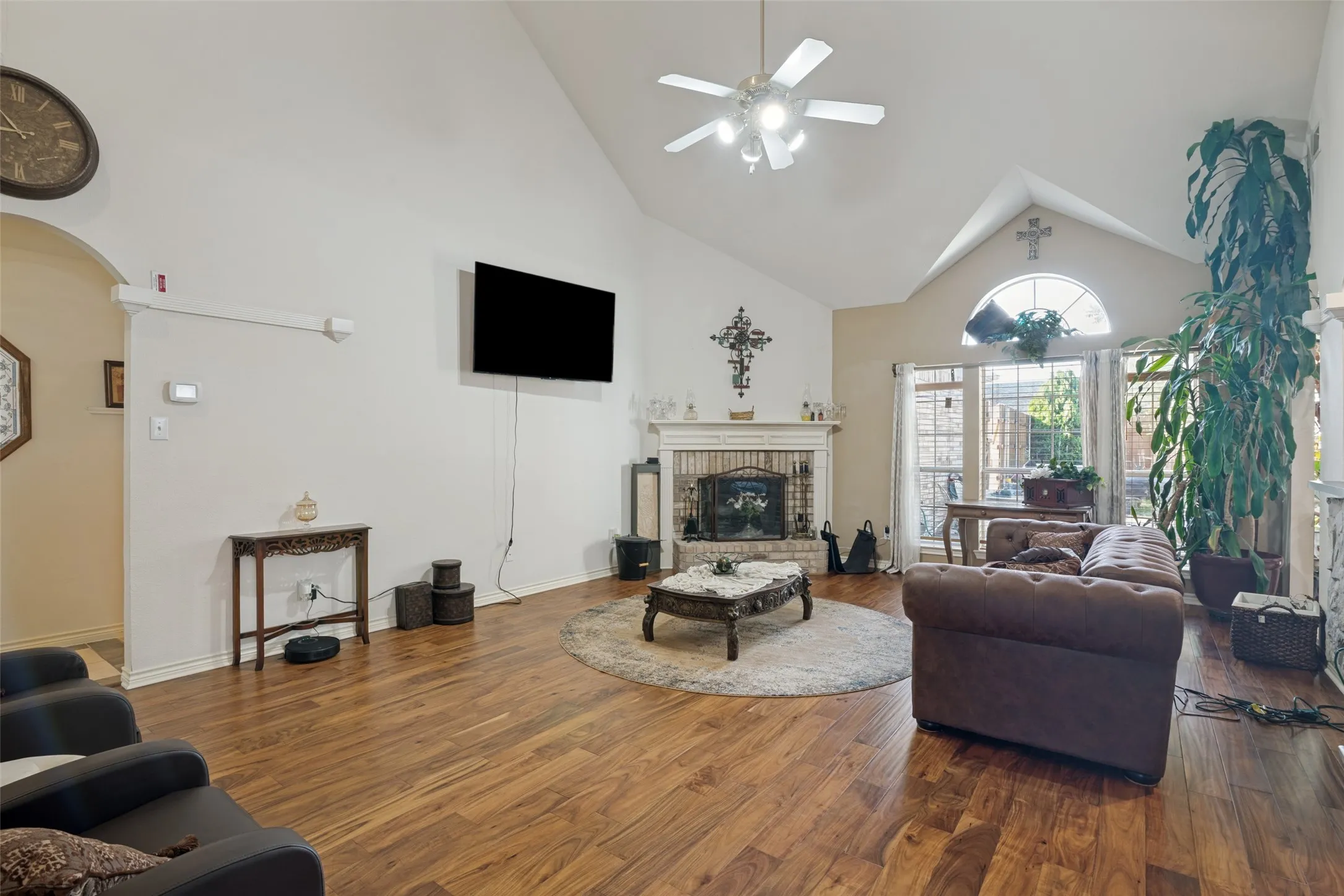 Vaulted ceiling with ceiling fan, wood-finish flooring, brick fireplace with mantel, arched transom window, and an arched entry