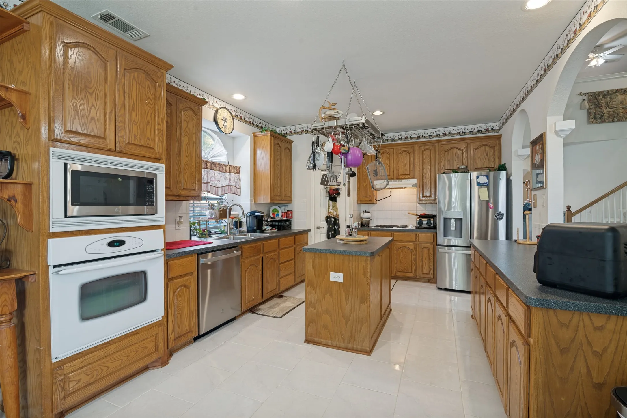 Kitchen with wood-finish cabinetry, white tile flooring, built-in stainless steel microwave and oven, and a central island