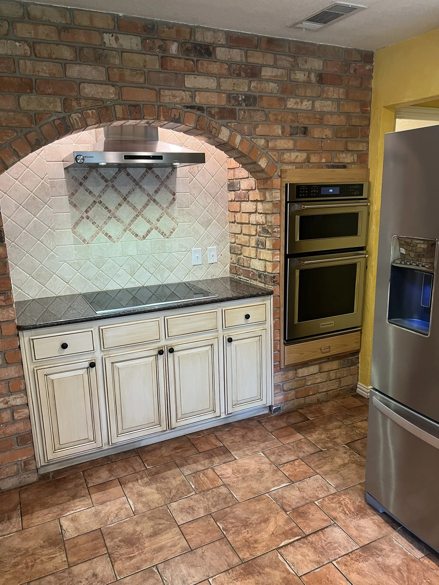 Exposed brick archway, stainless steel range hood, built-in double ovens, tile flooring, and light-colored cabinetry with dark hardware