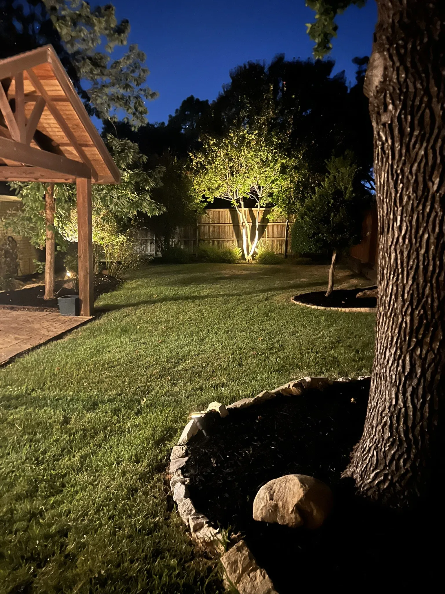 Expansive yard featuring a wooden pergola, mature trees, and a stone-edged planting bed