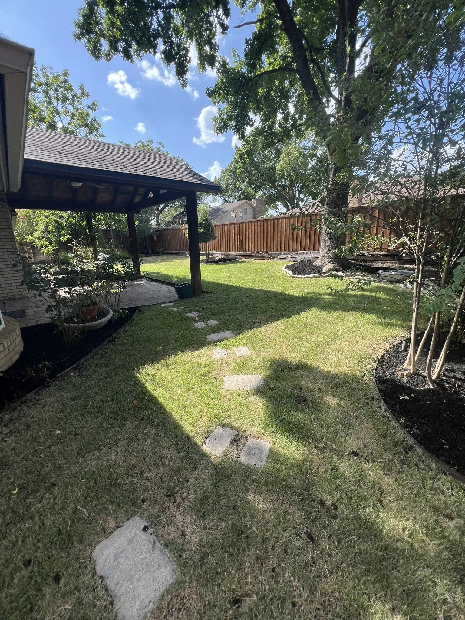 Expansive backyard featuring a covered patio, manicured lawn, stone paver pathway, mature trees, and a privacy fence